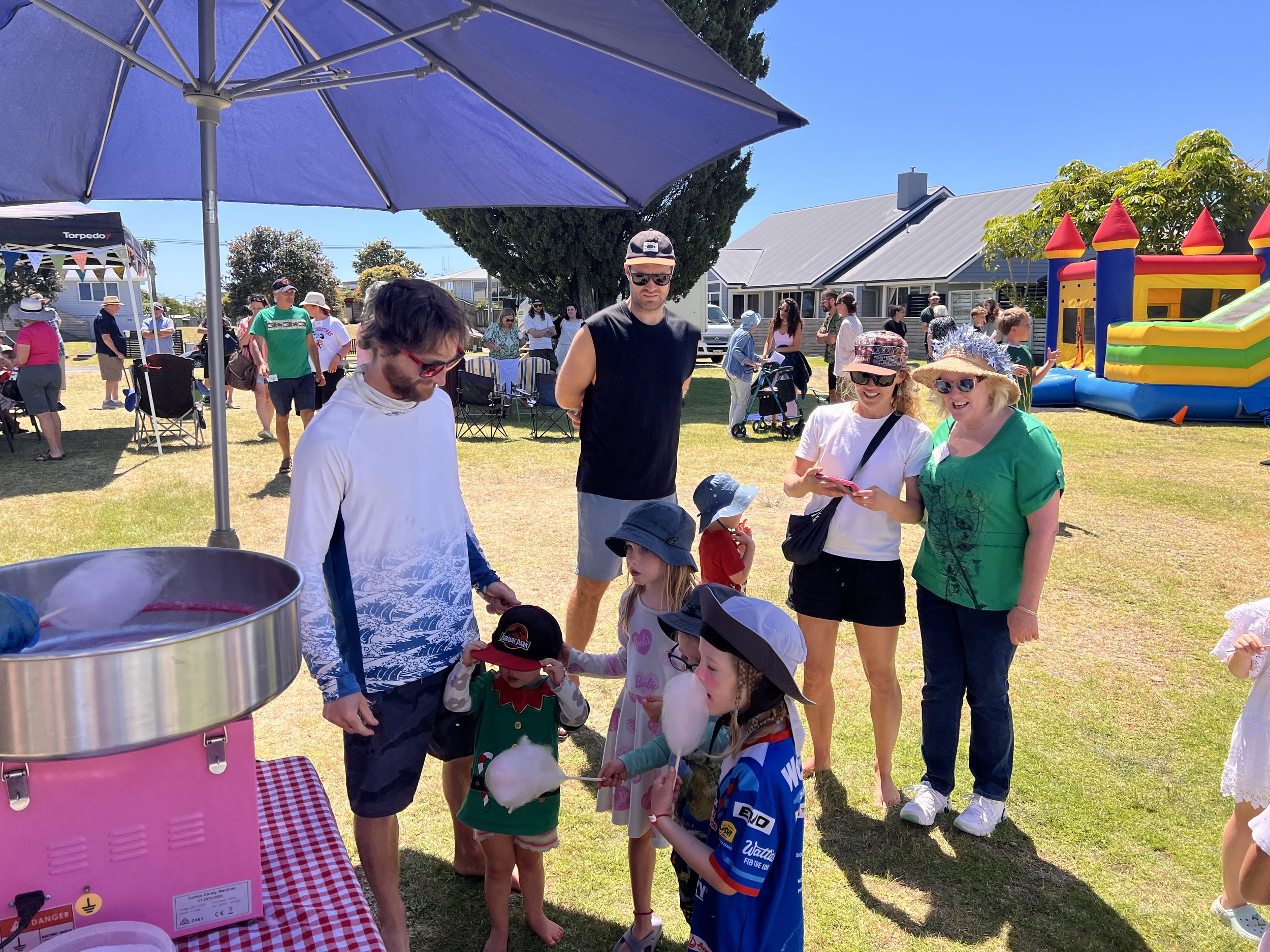 A group of people, including children and adults, waiting in line at an outdoor event on a sunny day. There is a bounce house in the background, and some people are wearing hats and sunglasses. The scene is set in a residential area with houses and trees visible.