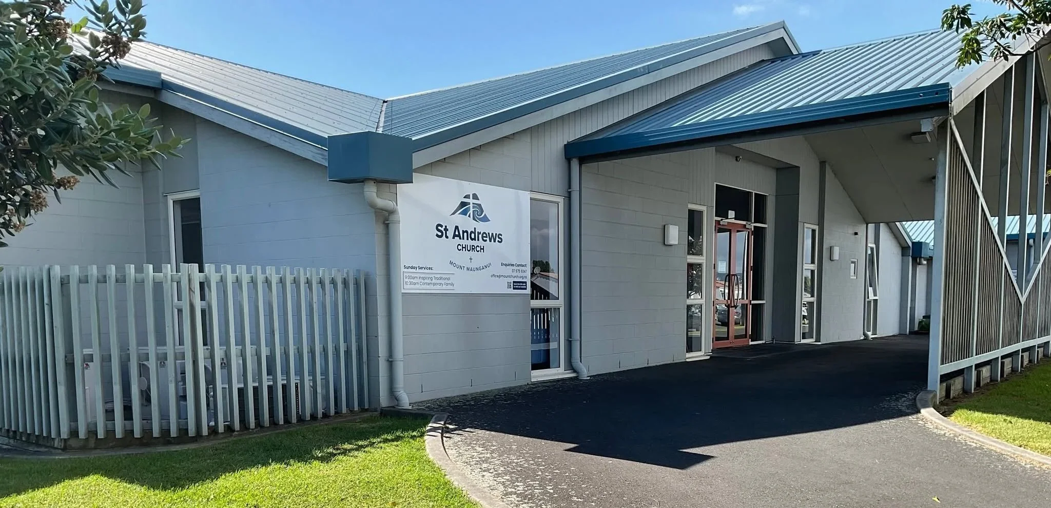 Exterior view of St Andrews Church in Mount Maunganui with a white fence, a pathway, and a sign displaying service times and contact information.
