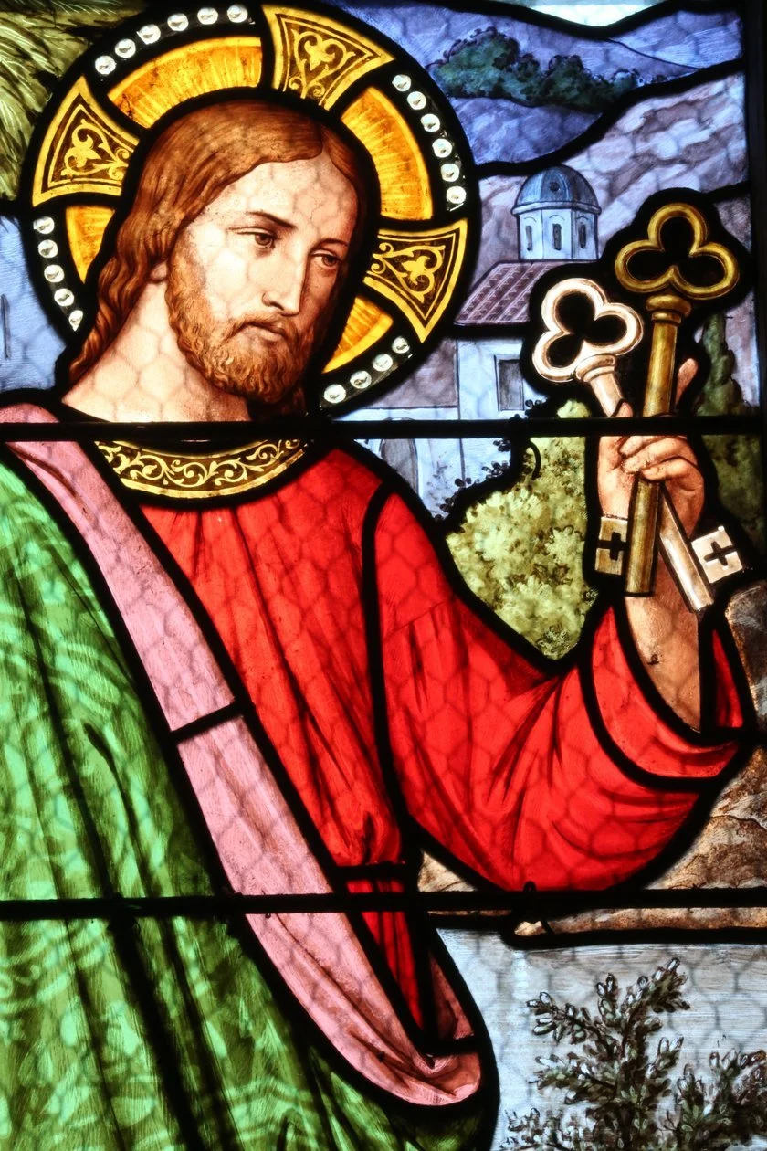 A stained glass window depicting Jesus Christ holding two keys, with a halo around his head, wearing a red robe and green cloak, with a church and trees in the background.