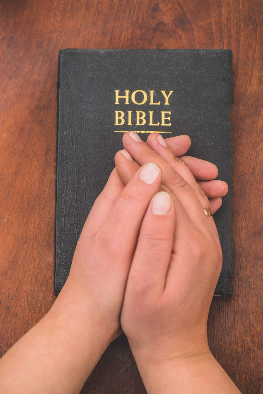 Hands clasped together resting on a closed black Holy Bible with gold lettering, on a wooden surface.