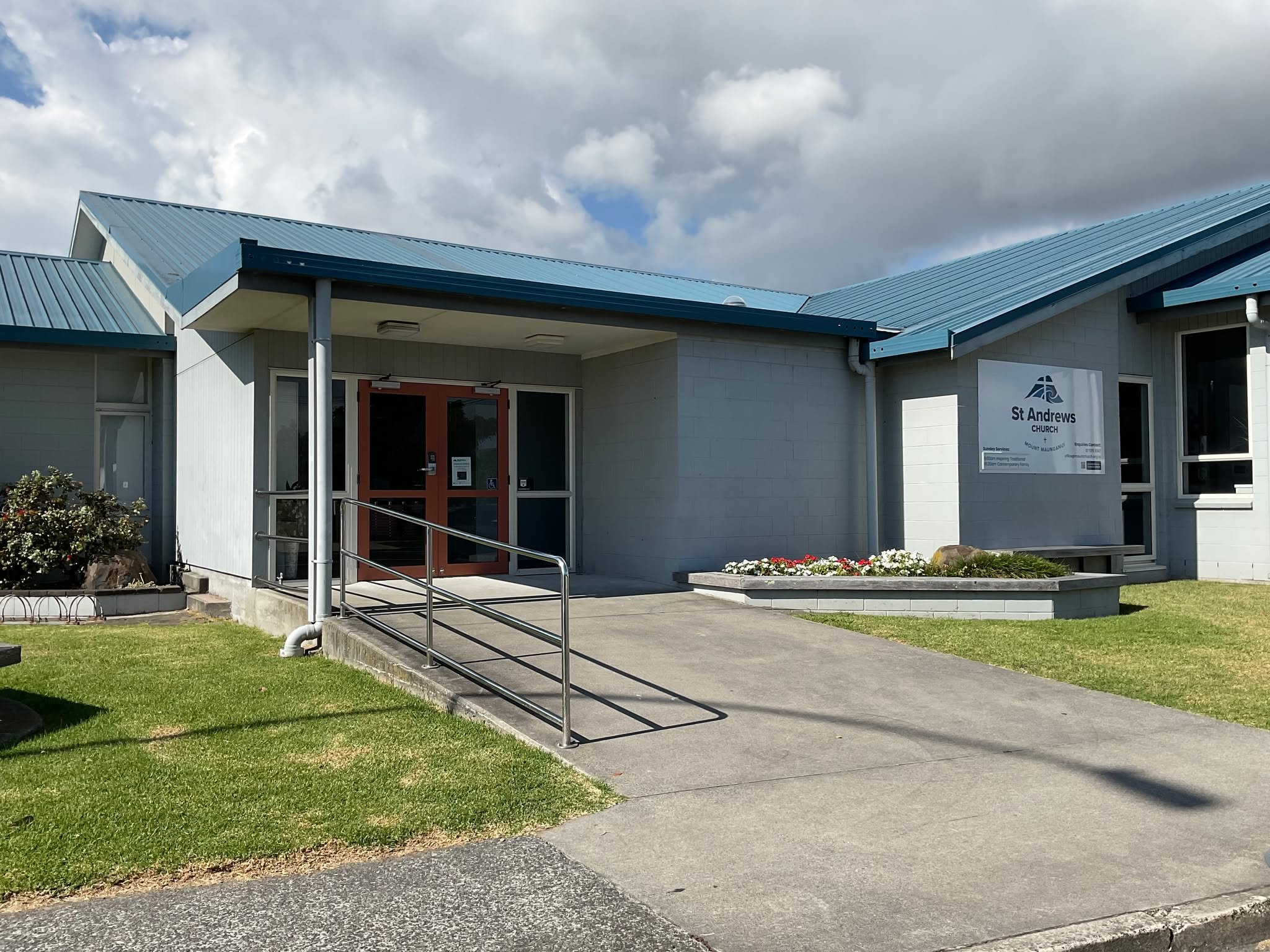 Exterior view of St Andrews Church building with a blue metal roof, a ramp for accessibility, a sign with the church's name, and well-maintained lawn and flowers in front.