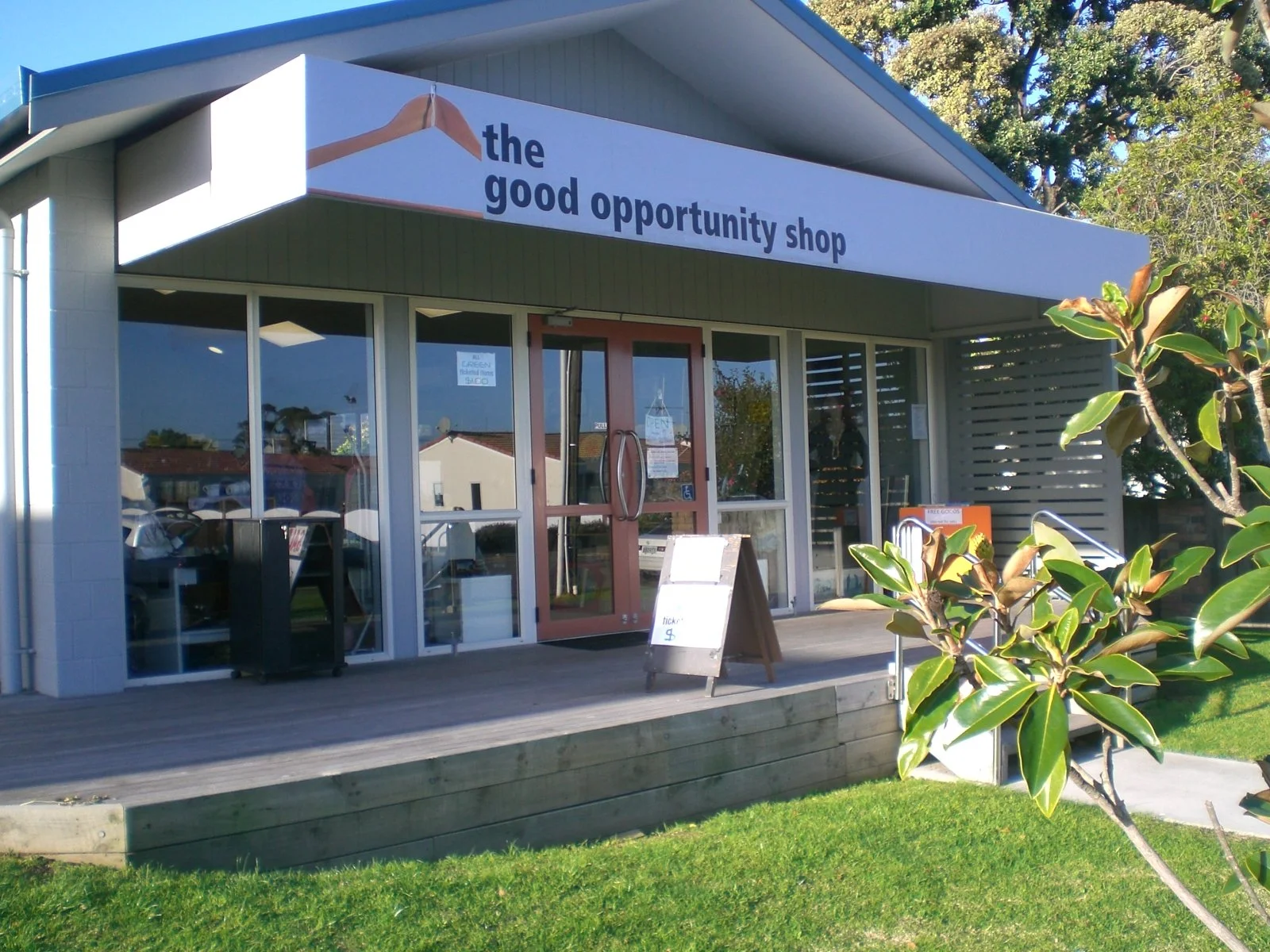 The storefront of 'The Good Opportunity Shop' with large glass windows, a red double door, a white signboard outside, and a sign overhead with a hand holding a pen. There are green plants in the foreground and clear skies overhead.