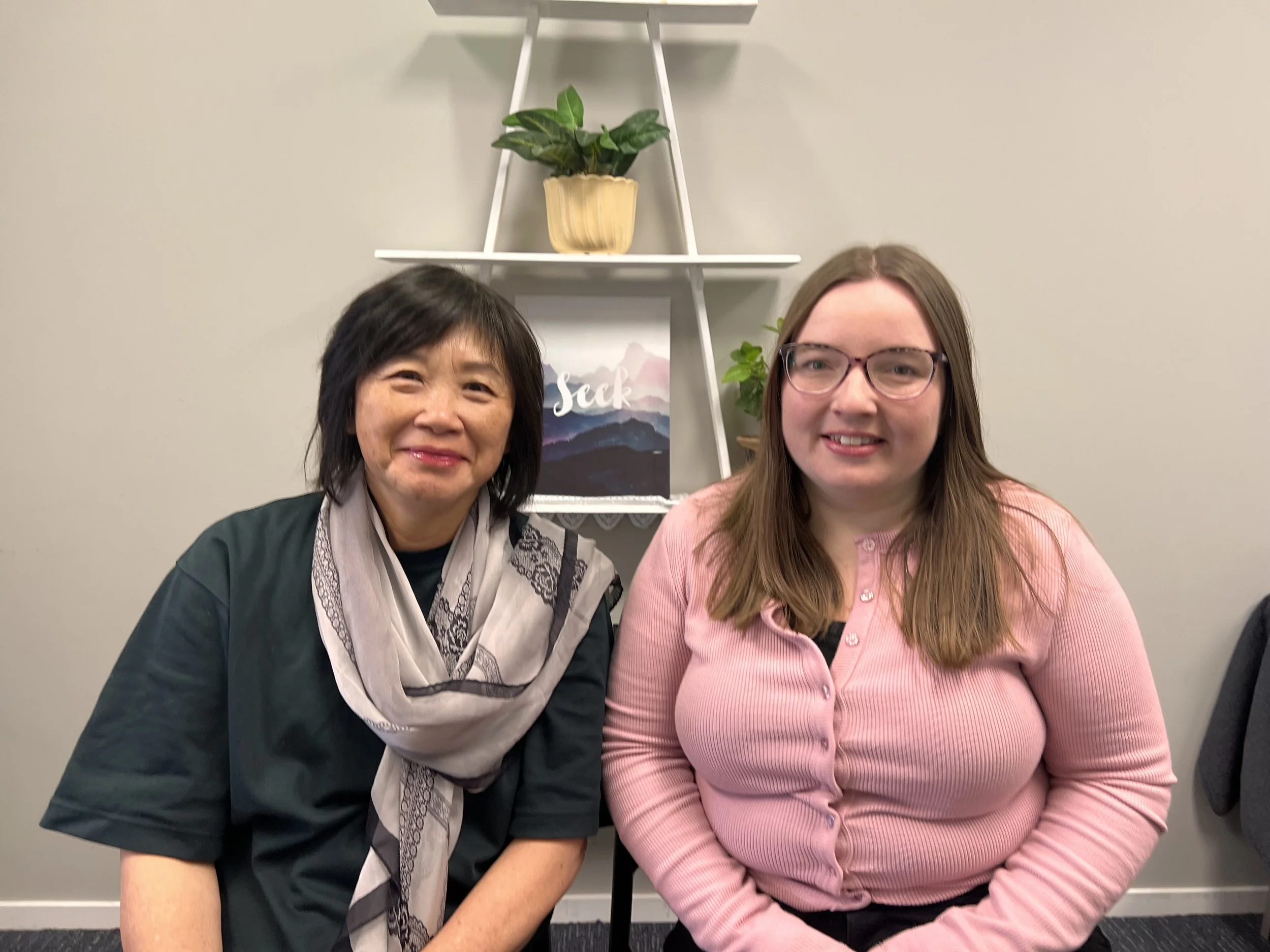 Two women sitting side by side in a room with a beige wall and white ladder-style shelf with potted plants and a framed picture. The woman on the left has dark hair, Asian features, and is wearing a dark top with a patterned scarf. Both women are smiling at the camera.