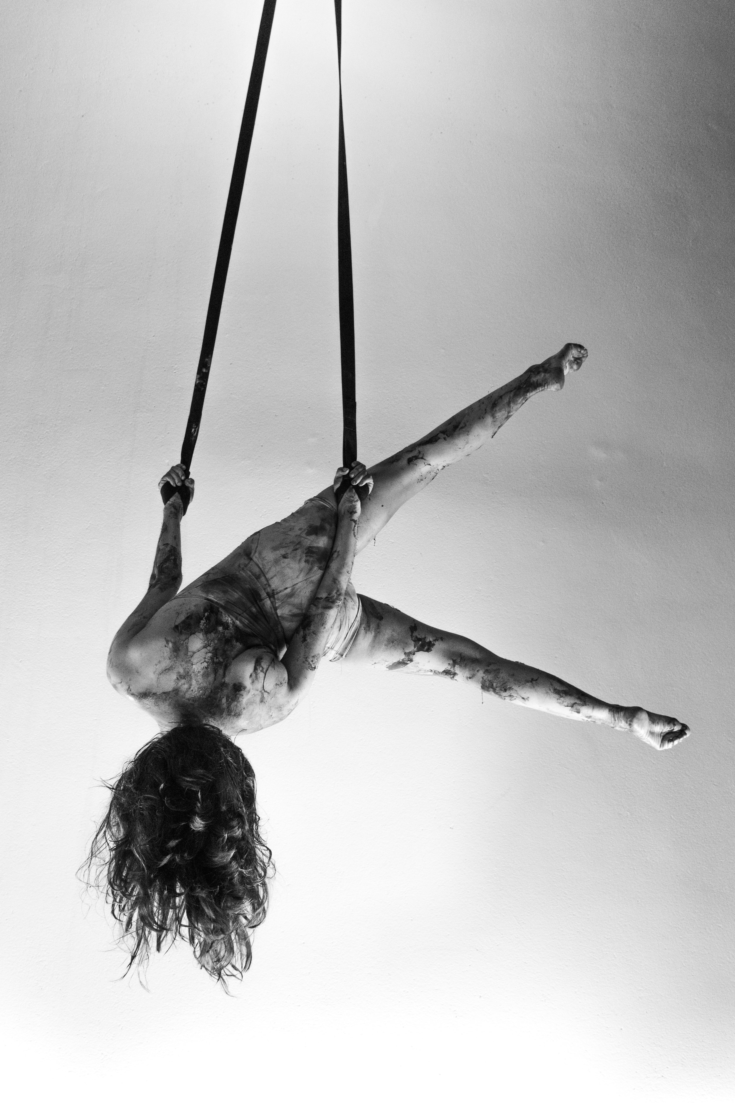 Una escultura de una mujer en una postura acrobática, colgada hacia abajo con las piernas extendidas hacia un lado, sosteniéndose con las manos en unas cuerdas.