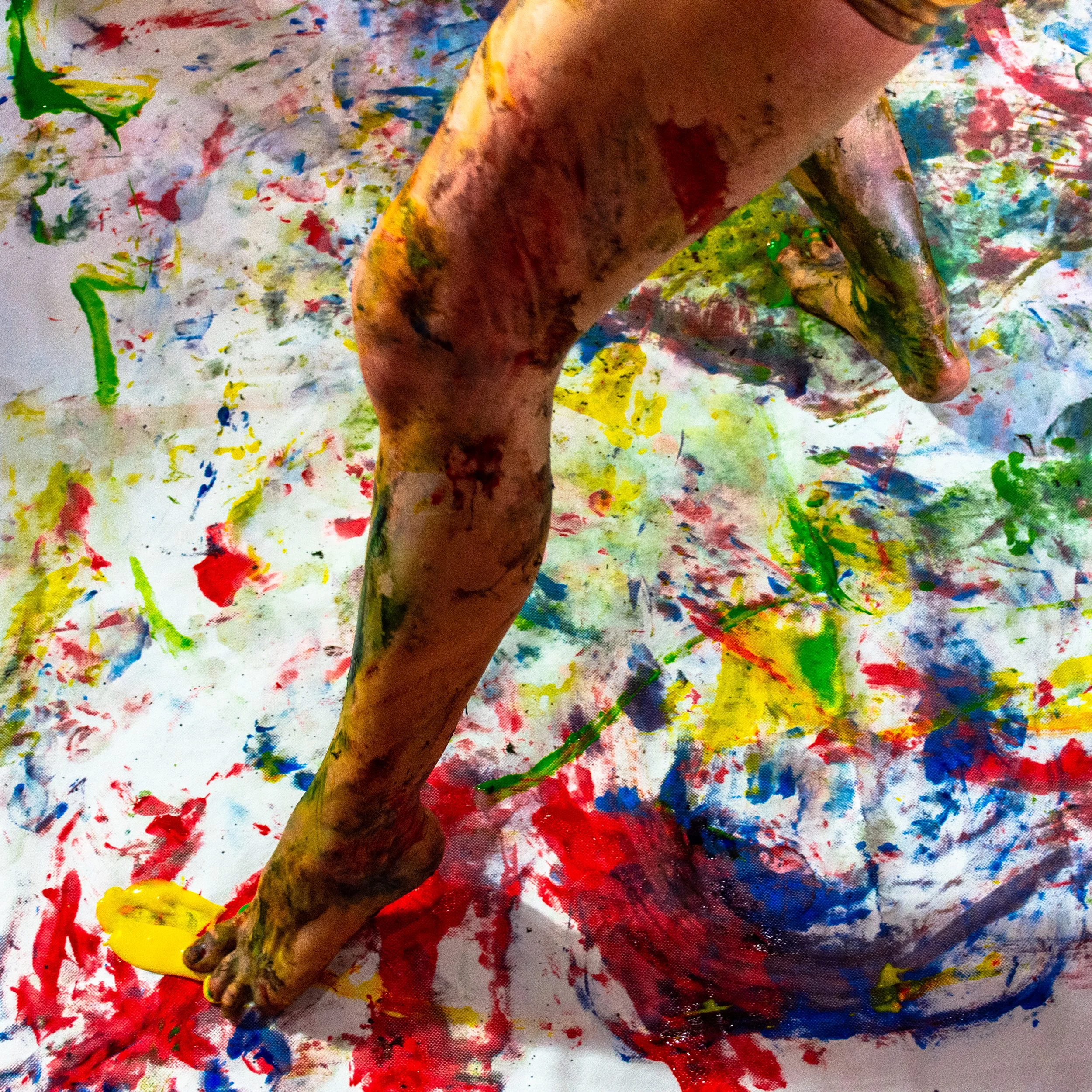 Mano cubierta de pintura de diversos colores, con piso de papel pintado también manchado de pintura, en un entorno artístico. 