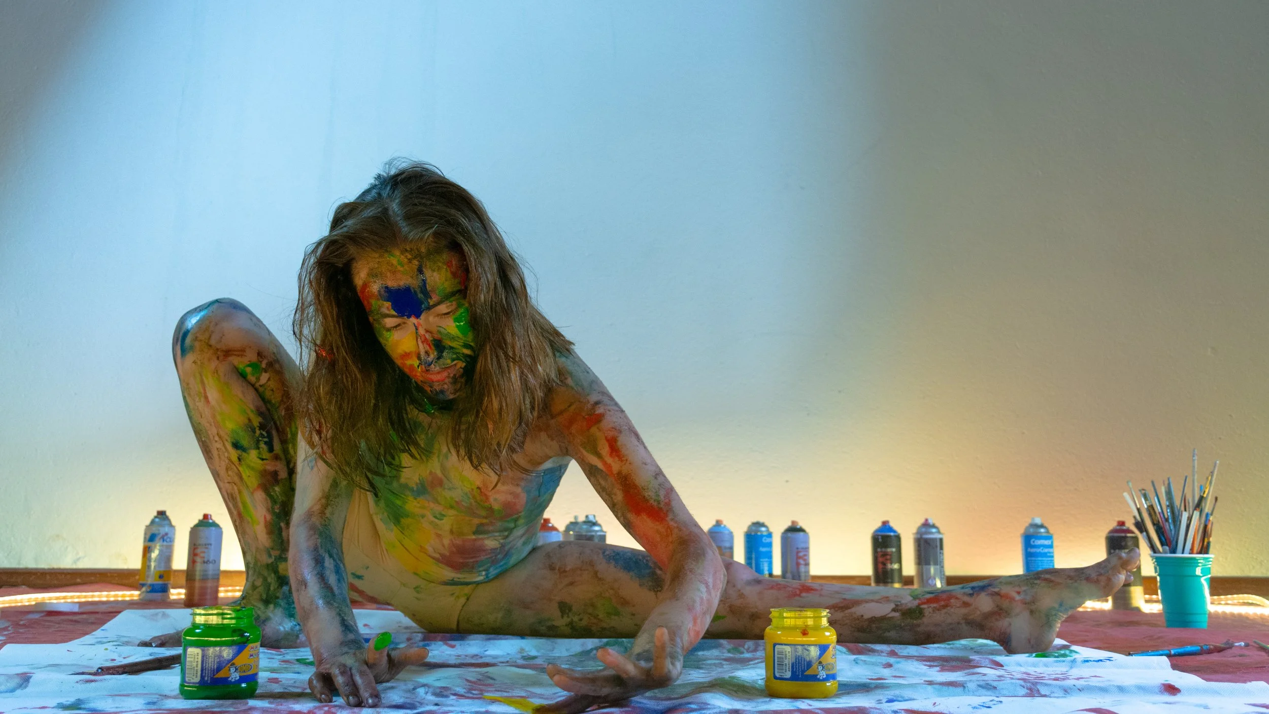 Mujer en posición de yoga cubierta de pintura de colores, rodeada de latas de pintura y pinceles en un ambiente artístico.