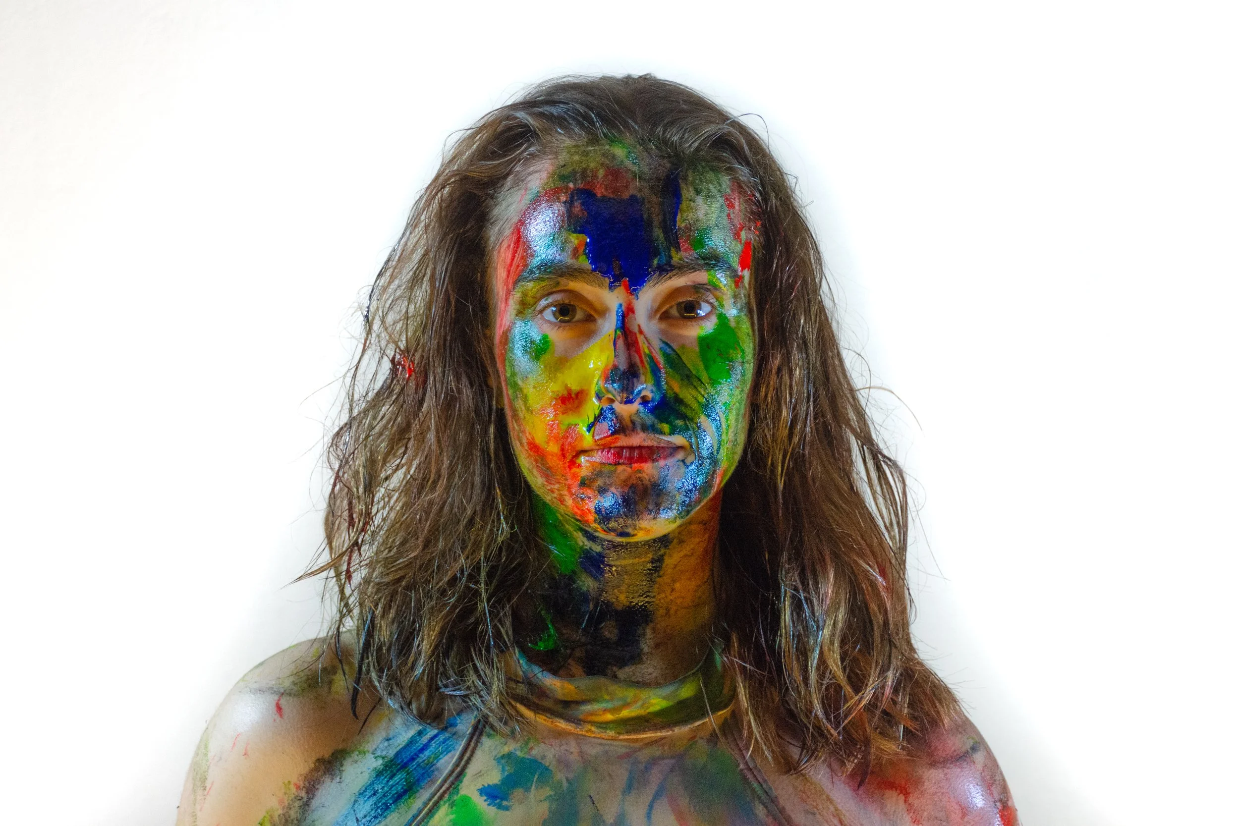 Mujer con cabello largo y rizado, con la cara y parte del cuerpo pintados con colores vivos y diversos, sobre fondo blanco.