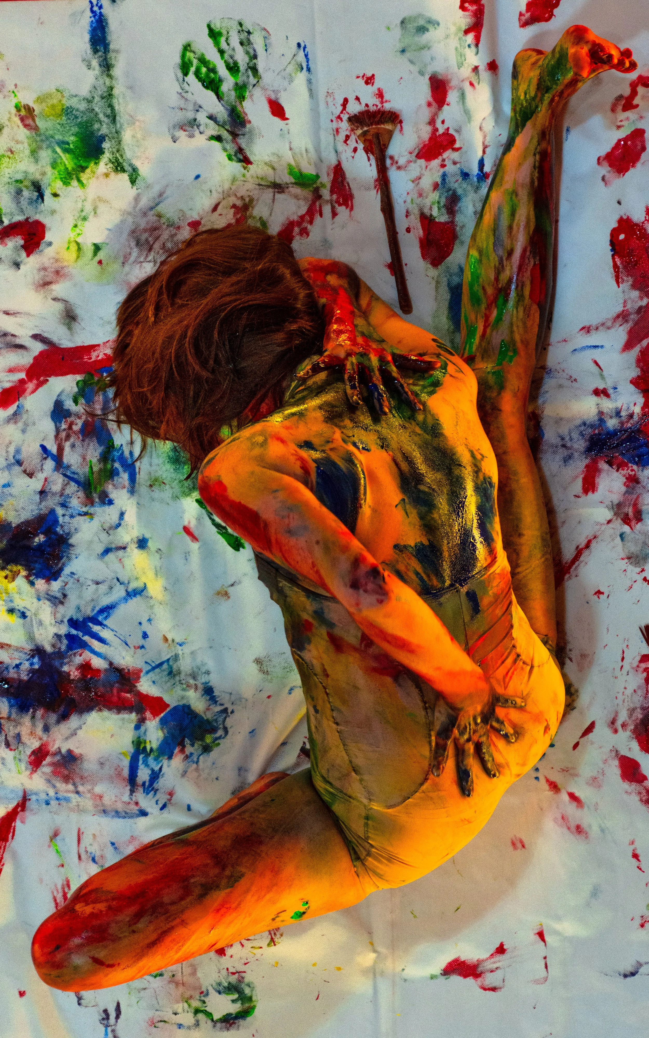 Persona pintando su cuerpo con colores vivos, rodeada de manchas de pintura en el suelo y la pared.