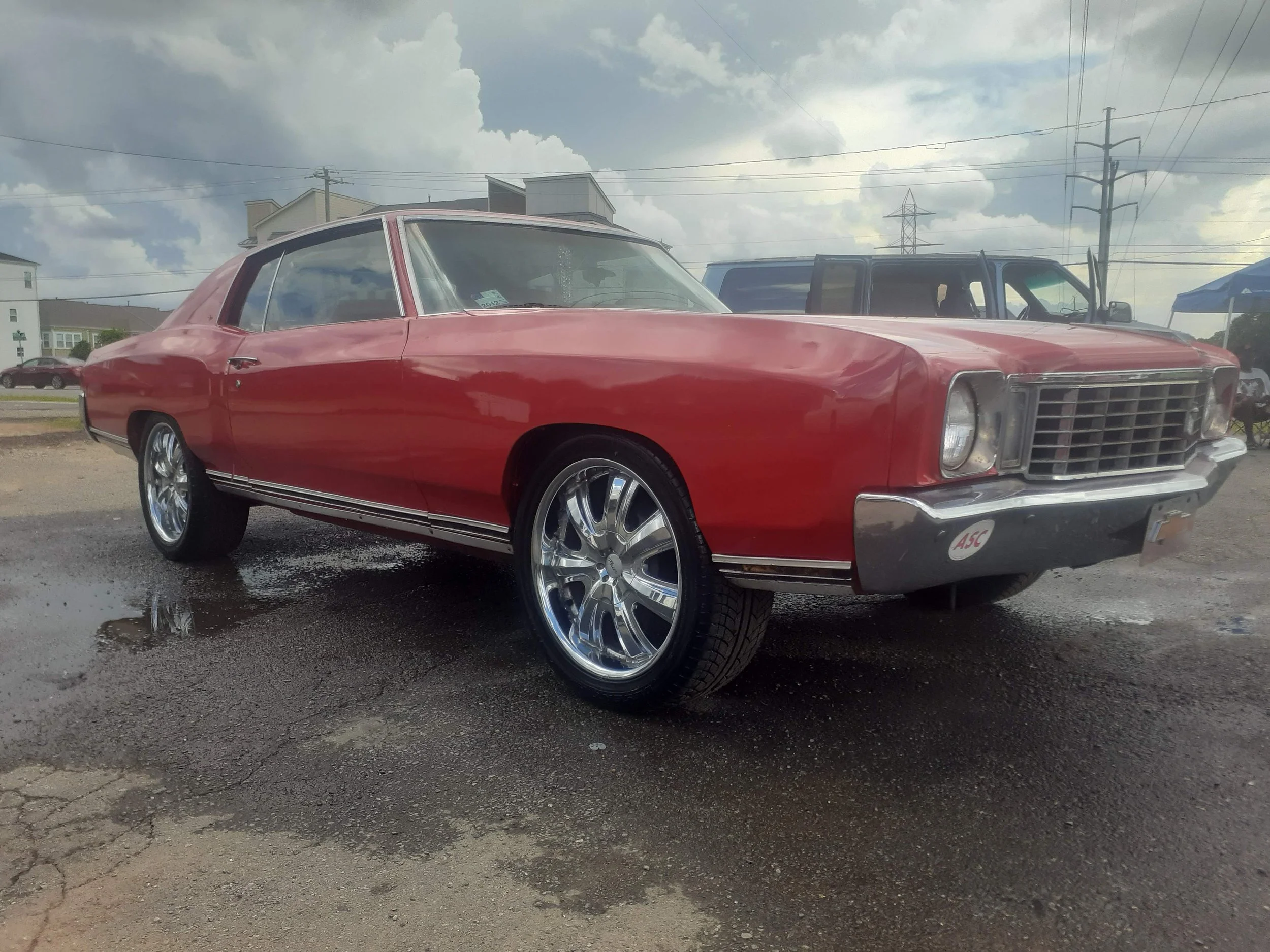 A vintage red car with shiny chrome wheels parked on a wet patch of asphalt in an outdoor lot. Cloudy sky with grey clouds overhead, power lines in the background, and other vehicles and buildings visible.