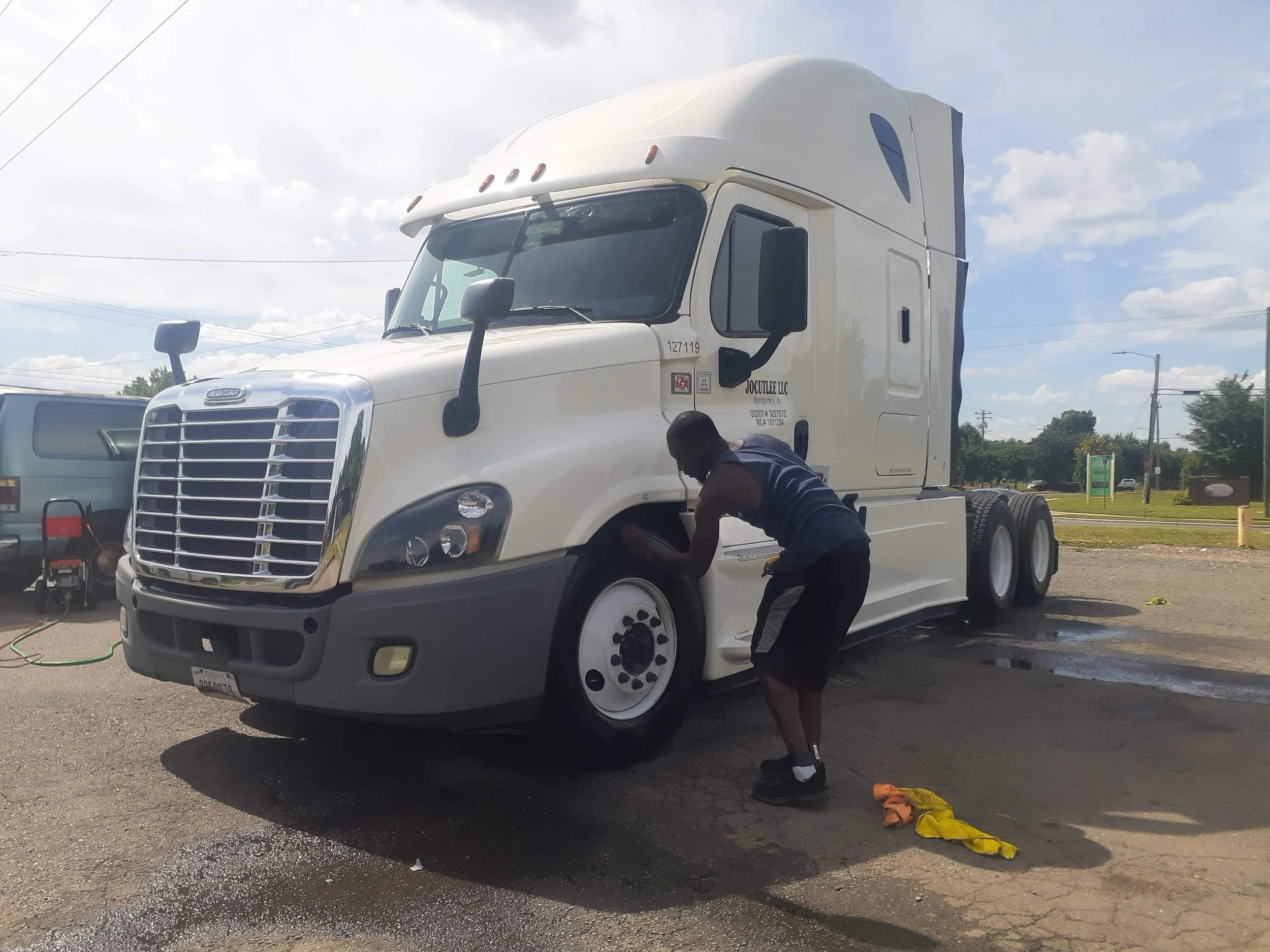 A man working on a white semi-truck in an outdoor parking lot, with a person leaning into the truck's engine area and tools and yellow cleaning cloth on the ground.