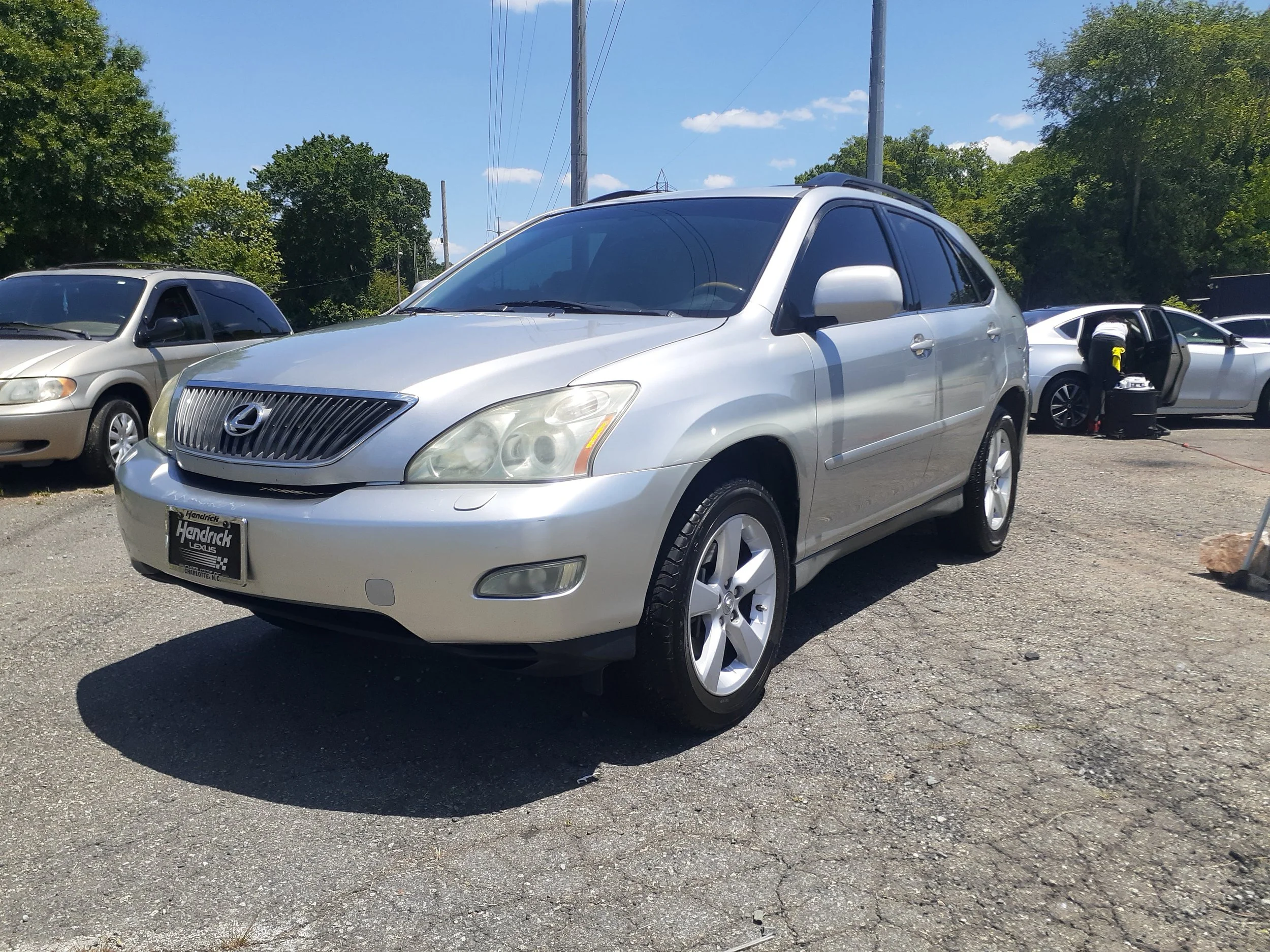 A silver Lexus SUV parked on a gravel lot with trees and utility poles in the background. The car has tinted windows and a dealership license plate frame from Hendrick Lexus.