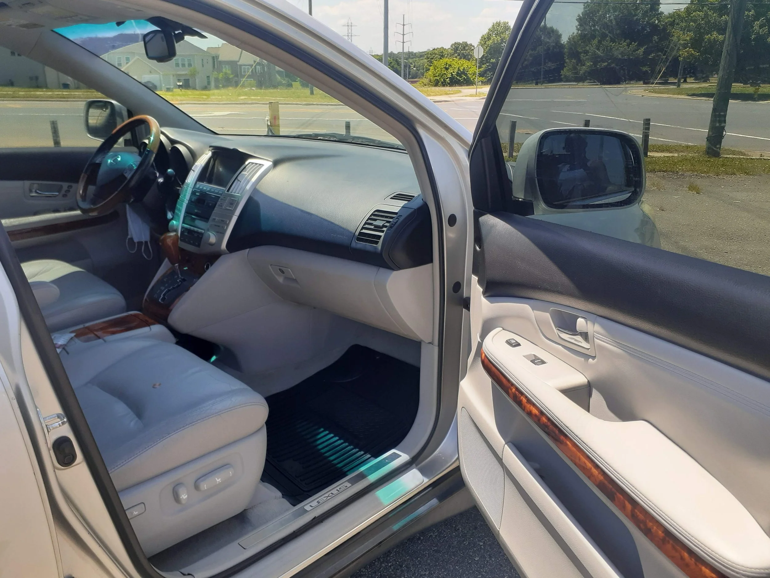 Inside view of a white SUV showing the driver and front passenger seats, dashboard, and side mirror; outside view of a parking lot with trees and houses.
