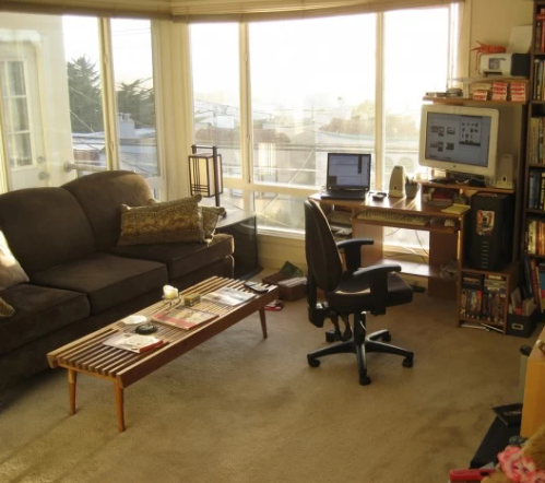 Living room with a large window, dark sofa, coffee table, desk with a computer, and an office chair.
