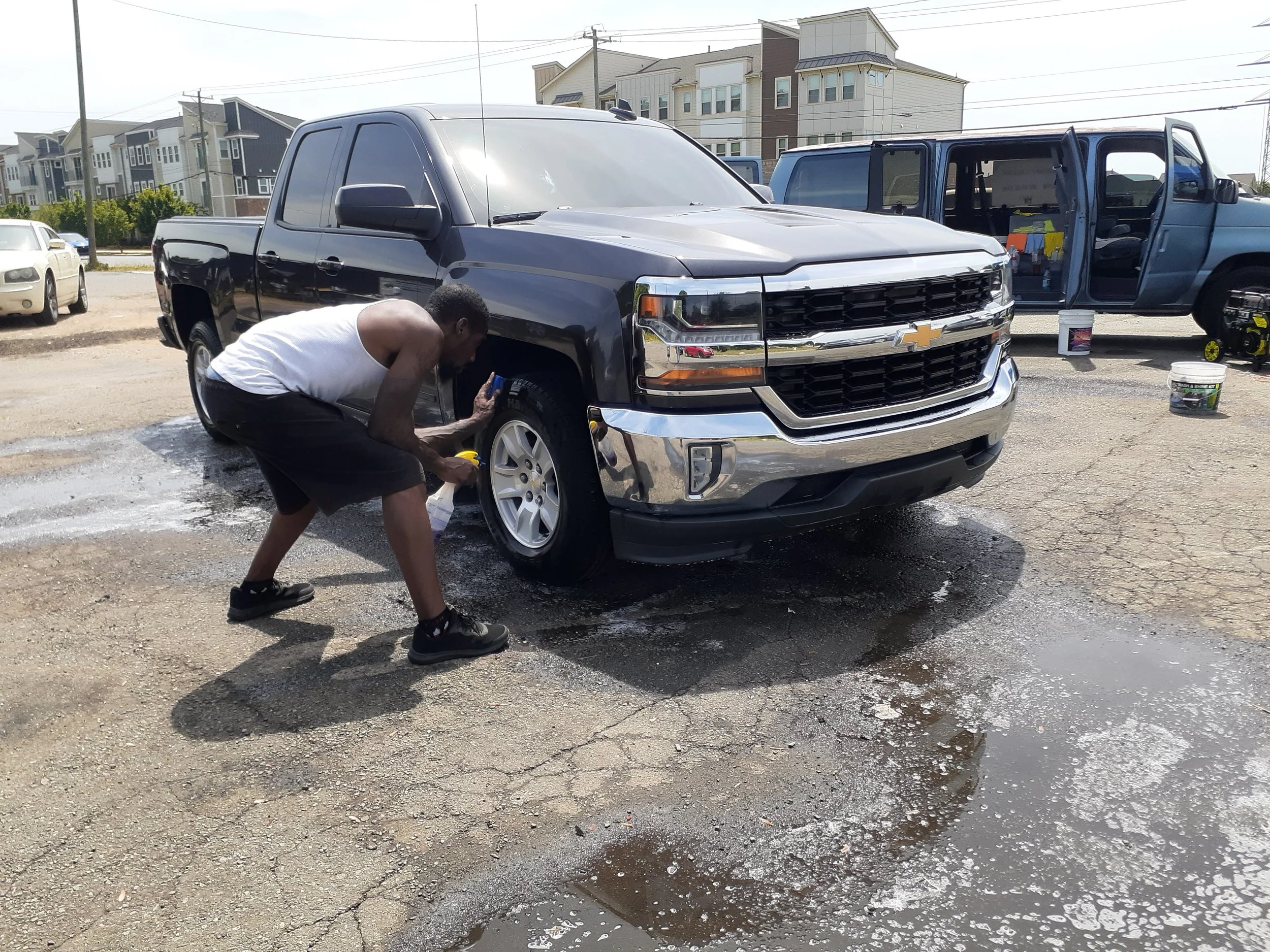 A man washing a black Chevrolet pickup truck with soap and water in an outdoor parking lot, with houses visible in the background.