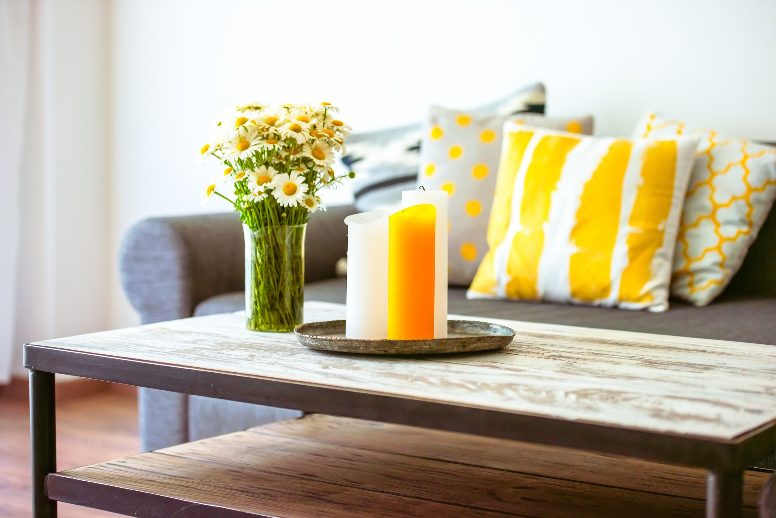 A wooden table with a silver tray holding three candles in white, pale yellow, and yellow, and a glass vase with a bouquet of daisies. Behind the table is a couch with decorative pillows in yellow, gray, and white patterns.