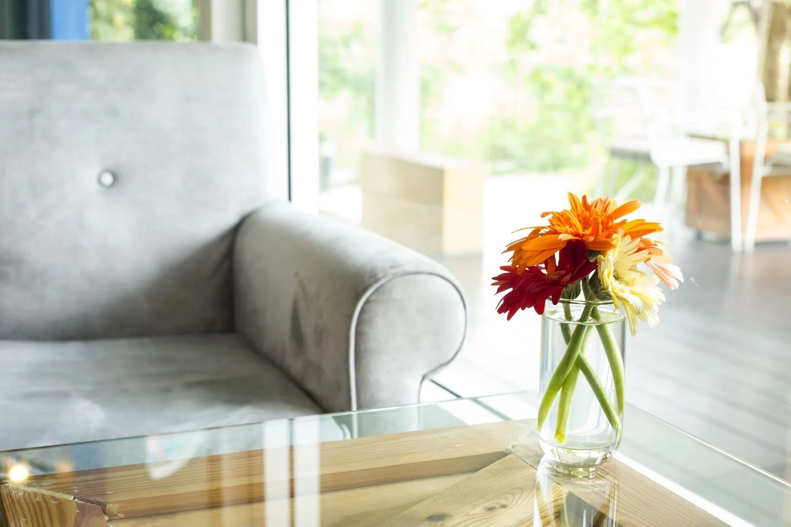 A glass vase with orange, red, yellow, and cream-colored flowers on a wooden table near a gray sofa, with a bright and airy room with large windows and outdoor seating in the background.