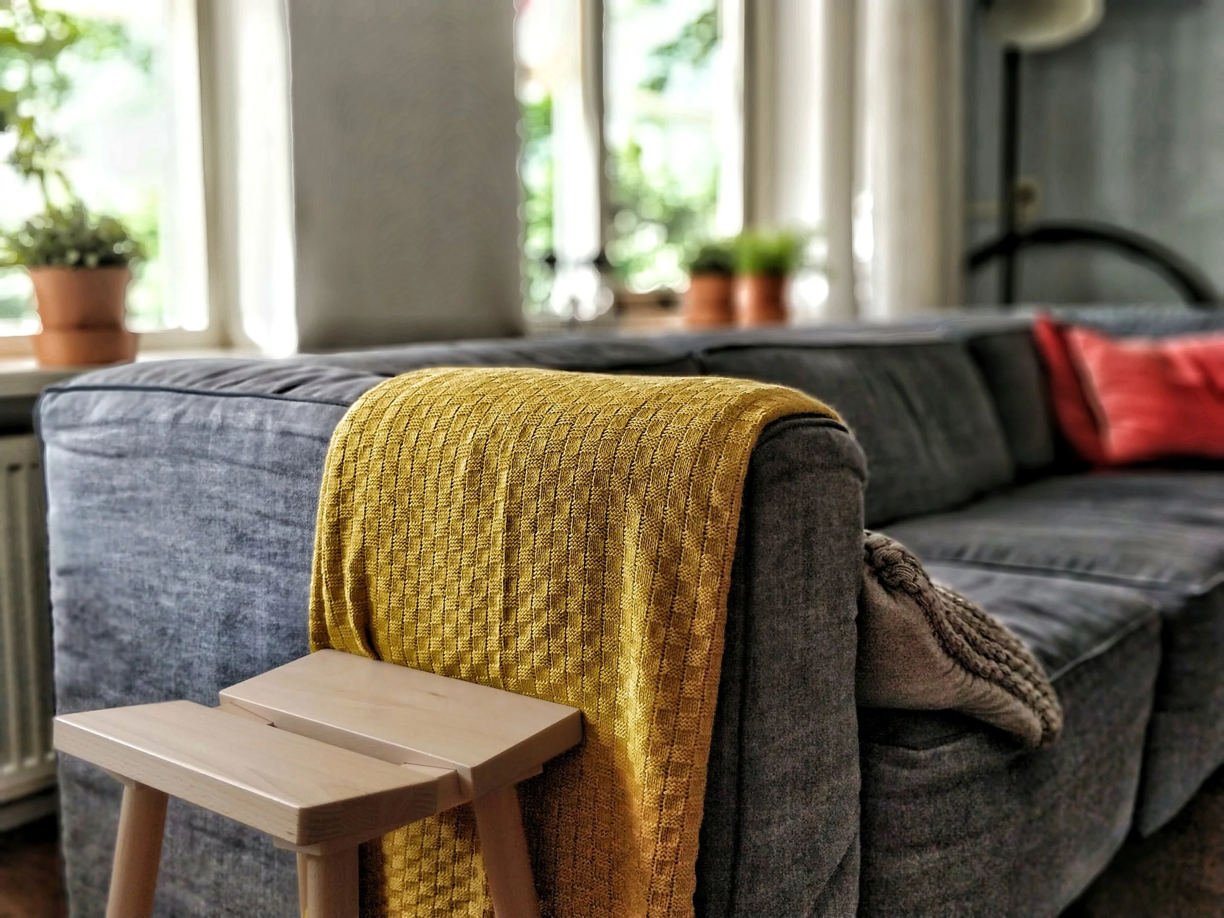 Close-up of a gray upholstered sofa with a yellow textured blanket draped over the armrest. In the background, there is a window with white curtains, potted plants on the windowsill, and blurred home interior elements.