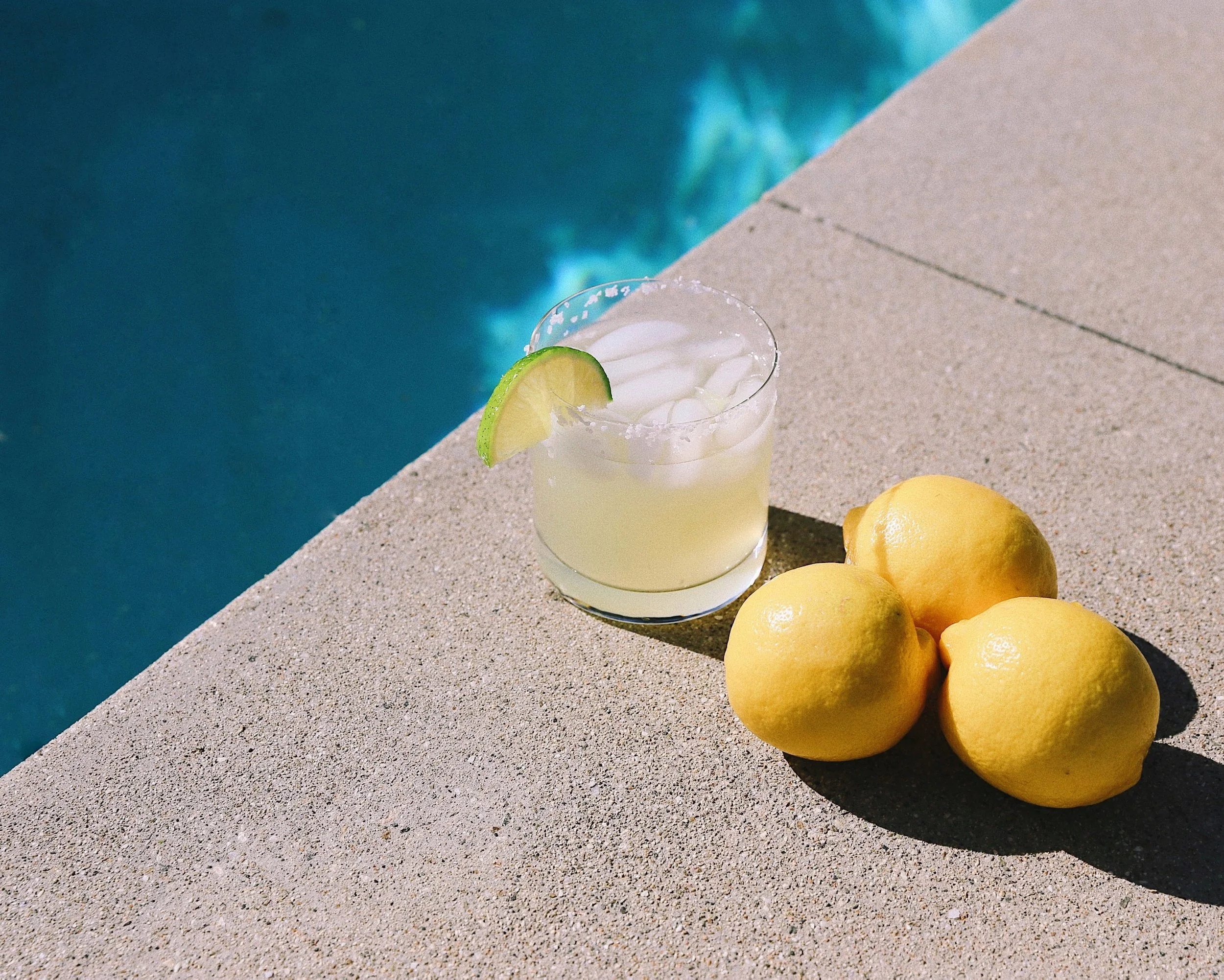 A close-up of a glass of lemonade with ice and a lime wedge on a concrete surface beside several whole lemons, with a swimming pool in the background.