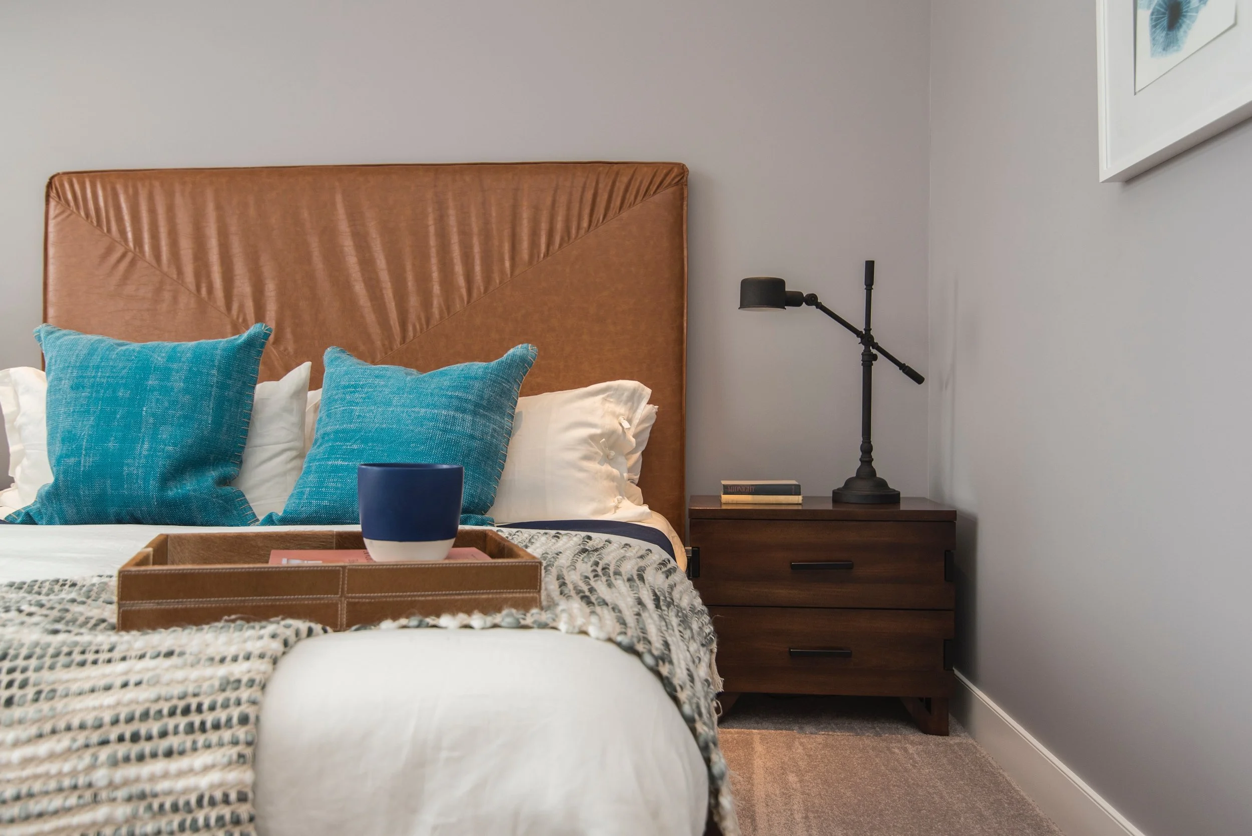 A bedroom with a large brown headboard, two blue pillows, a nightstand with a black lamp, and a wall painting.