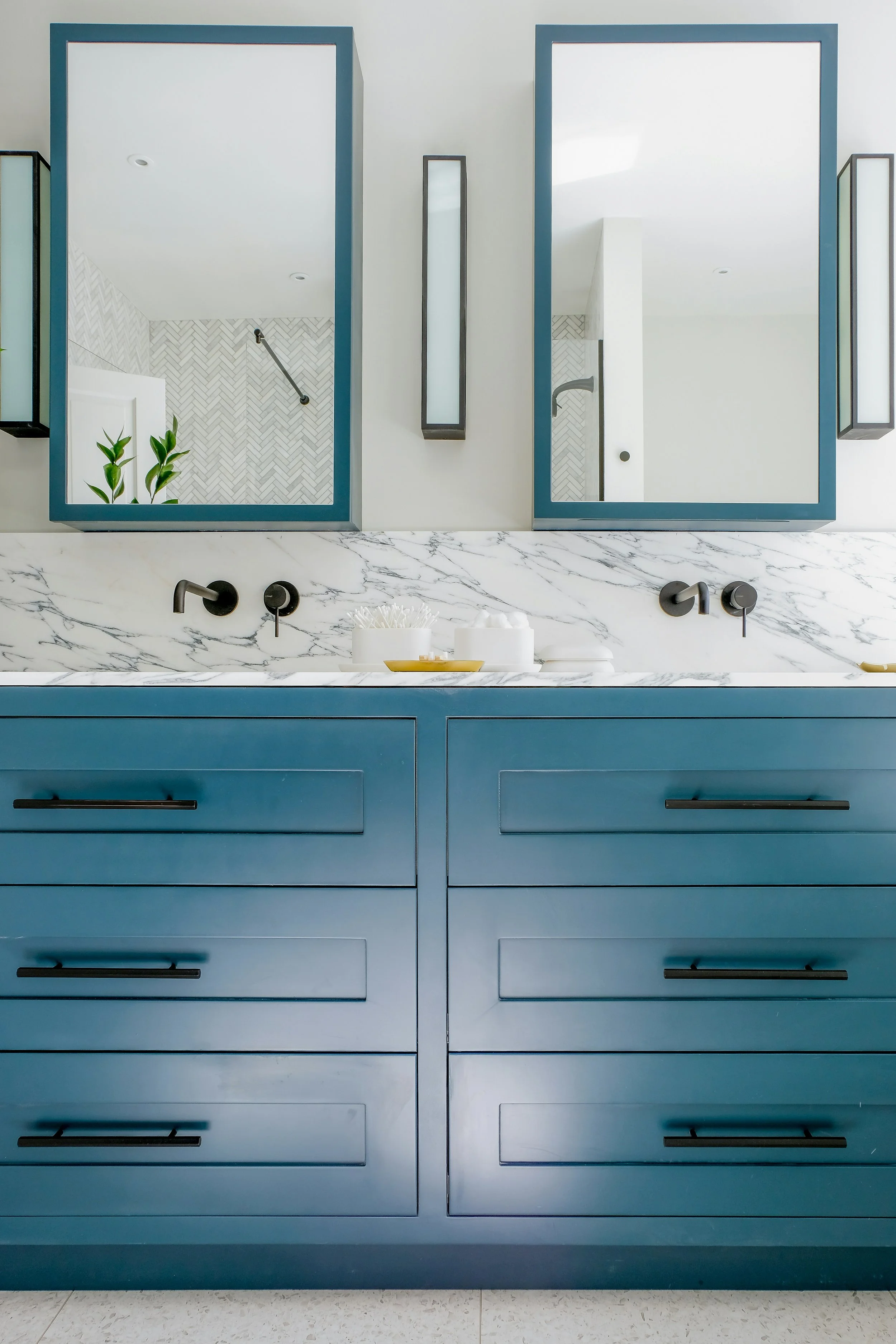Modern bathroom vanity with blue cabinets, marble countertop, and large mirror with black frames, reflecting a shower area with a herringbone tile wall and plants.