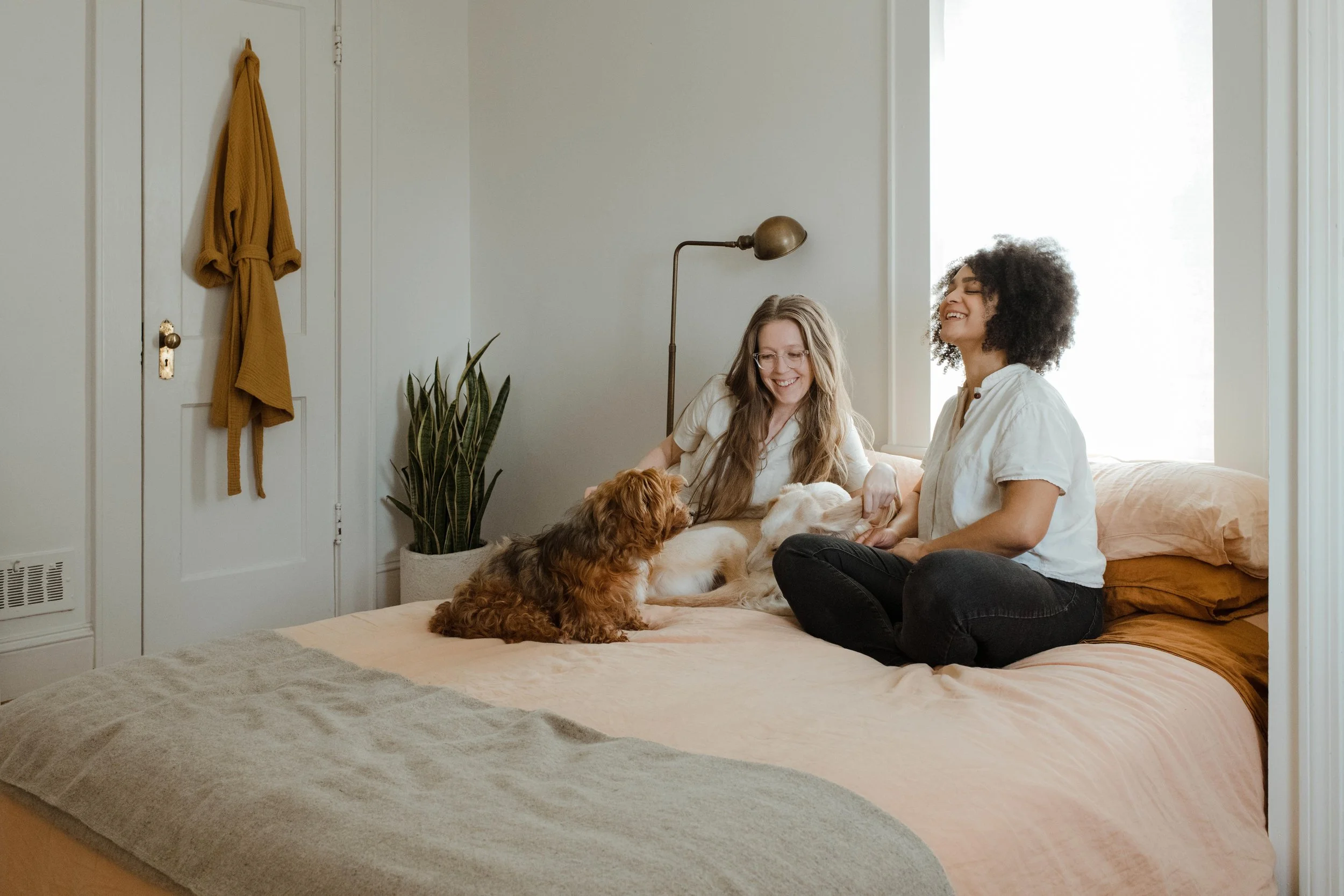 Two women sitting on a bed, smiling and playing with dogs in a bedroom with a large window, plant, and a yellow robe hanging on the door.