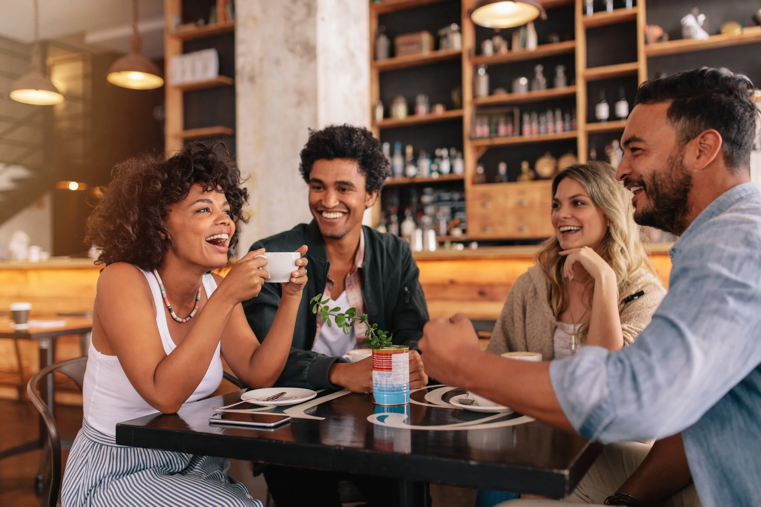 A group of four friends sitting at a table in a coffee shop, smiling and laughing while chatting. The woman on the left is holding a coffee mug, and there are various items on the table, including a smartphone and a can of drink. The background shows wooden shelves with bottles and jars, and warm lighting.