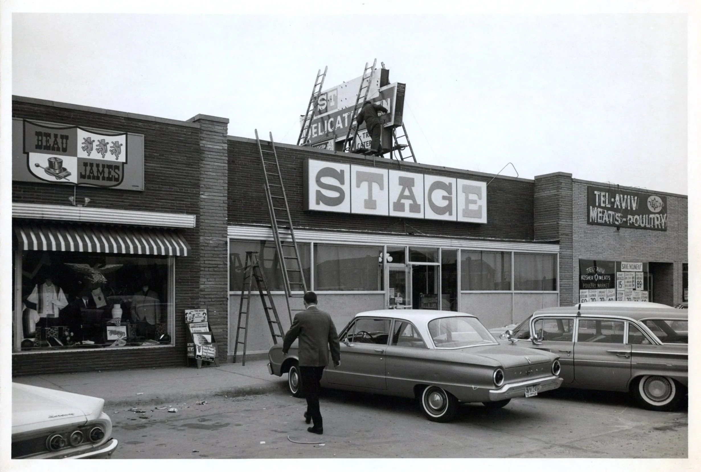 A black and white photo of a street scene showing storefronts and parked cars. A man is walking and another is working on a sign on the building. The sign reads 'STAGE'.