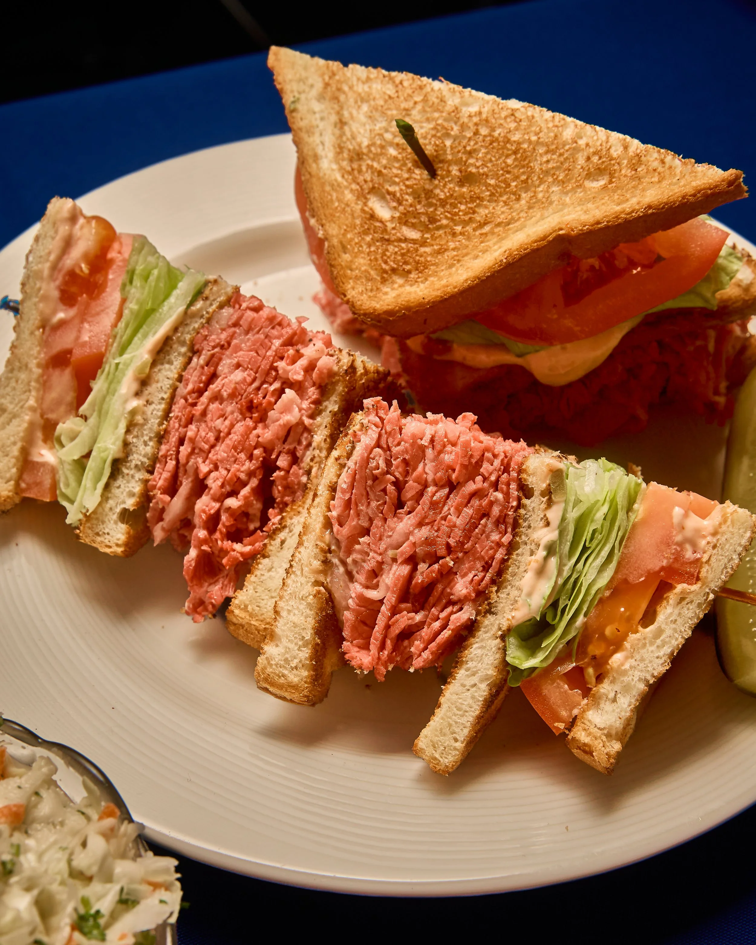 Close-up of a plate with a club sandwich featuring toasted bread, lettuce, tomato, and multiple types of deli meats, with a small side dish of potato salad in the corner.