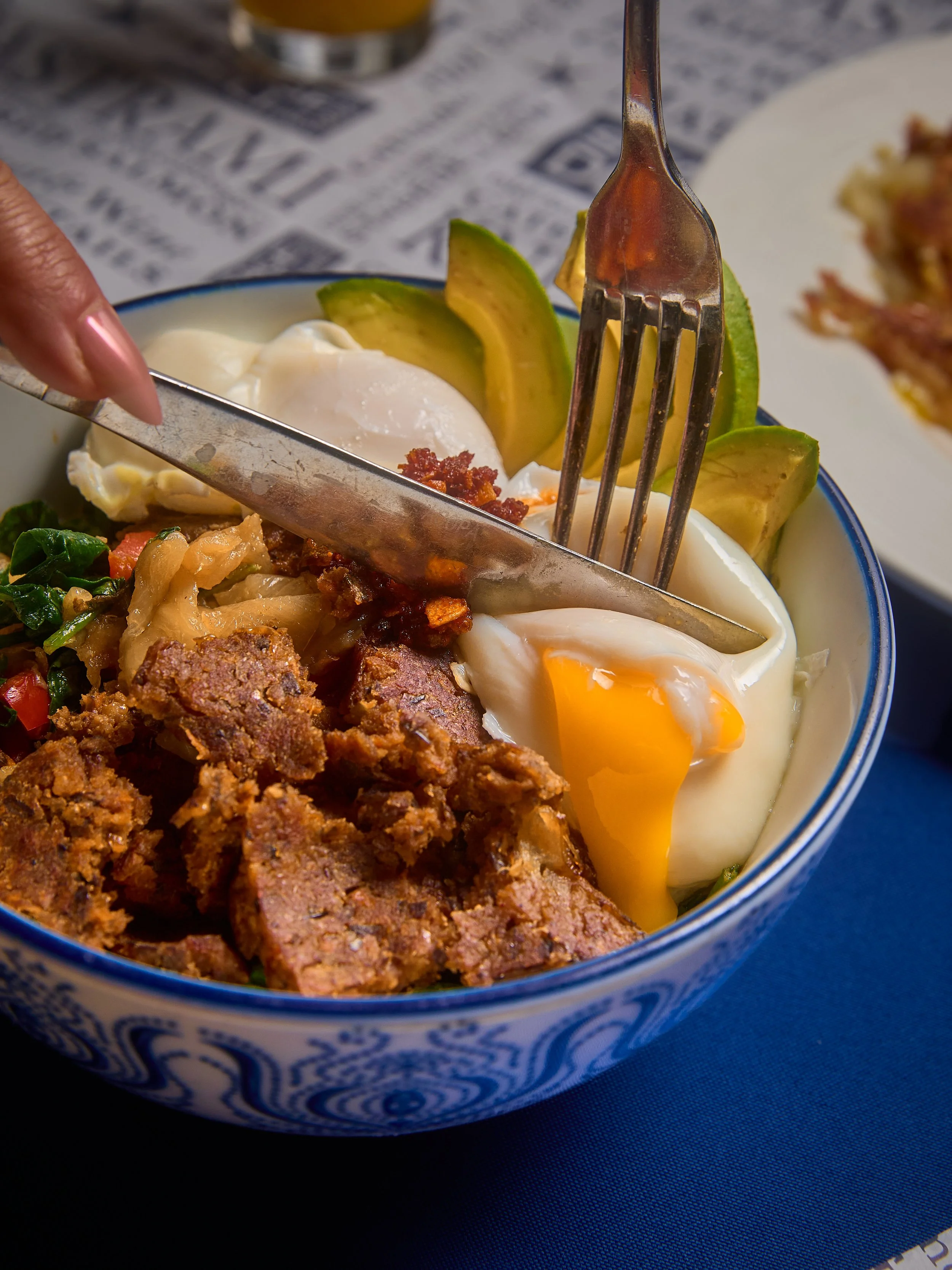 Close-up of a breakfast bowl containing fried egg, slices of avocado, cooked ground meat, and some cooked greens in a blue and white patterned bowl on a dark surface.