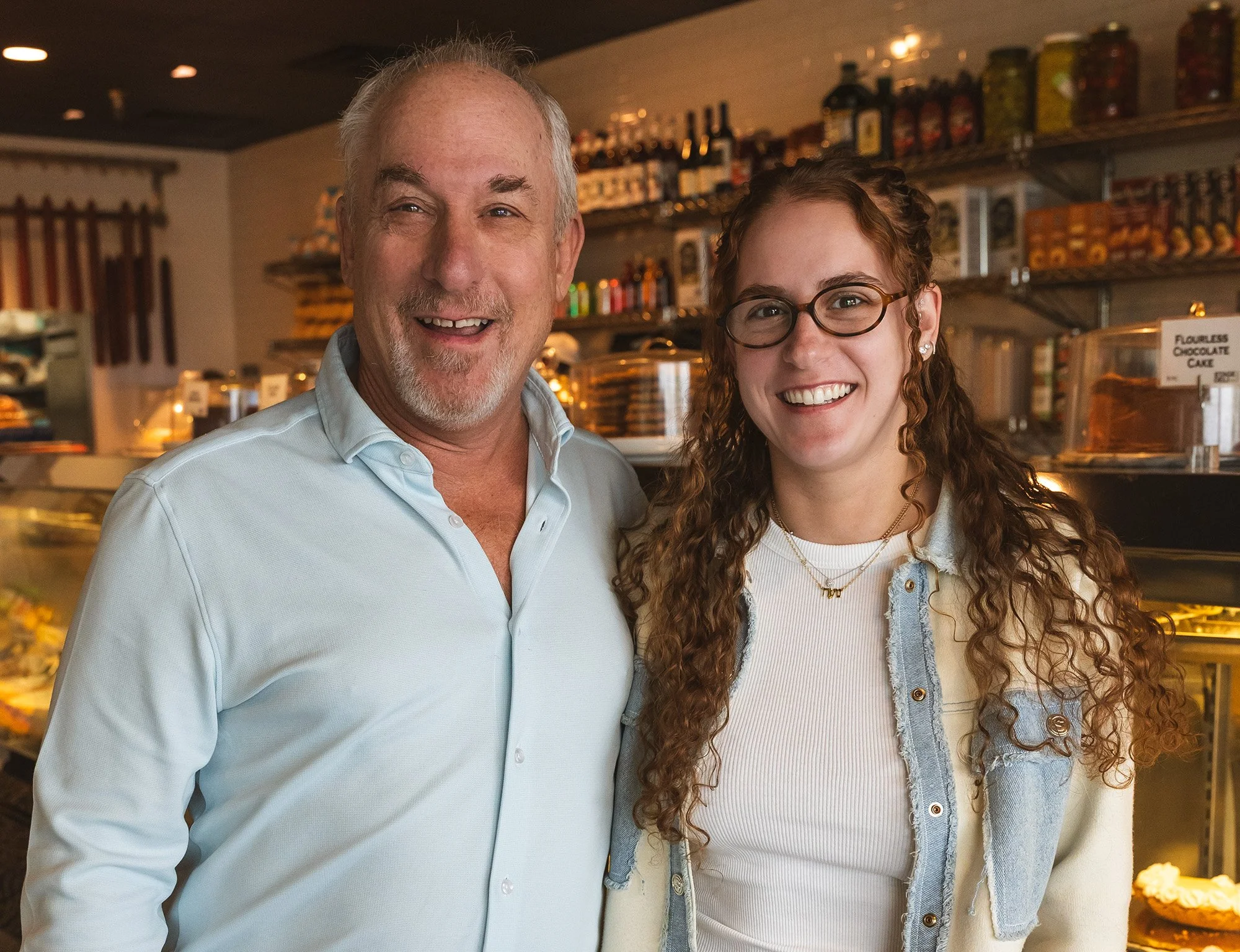 Two people smiling in a store or bakery, with shelves of jars and baked goods in the background.