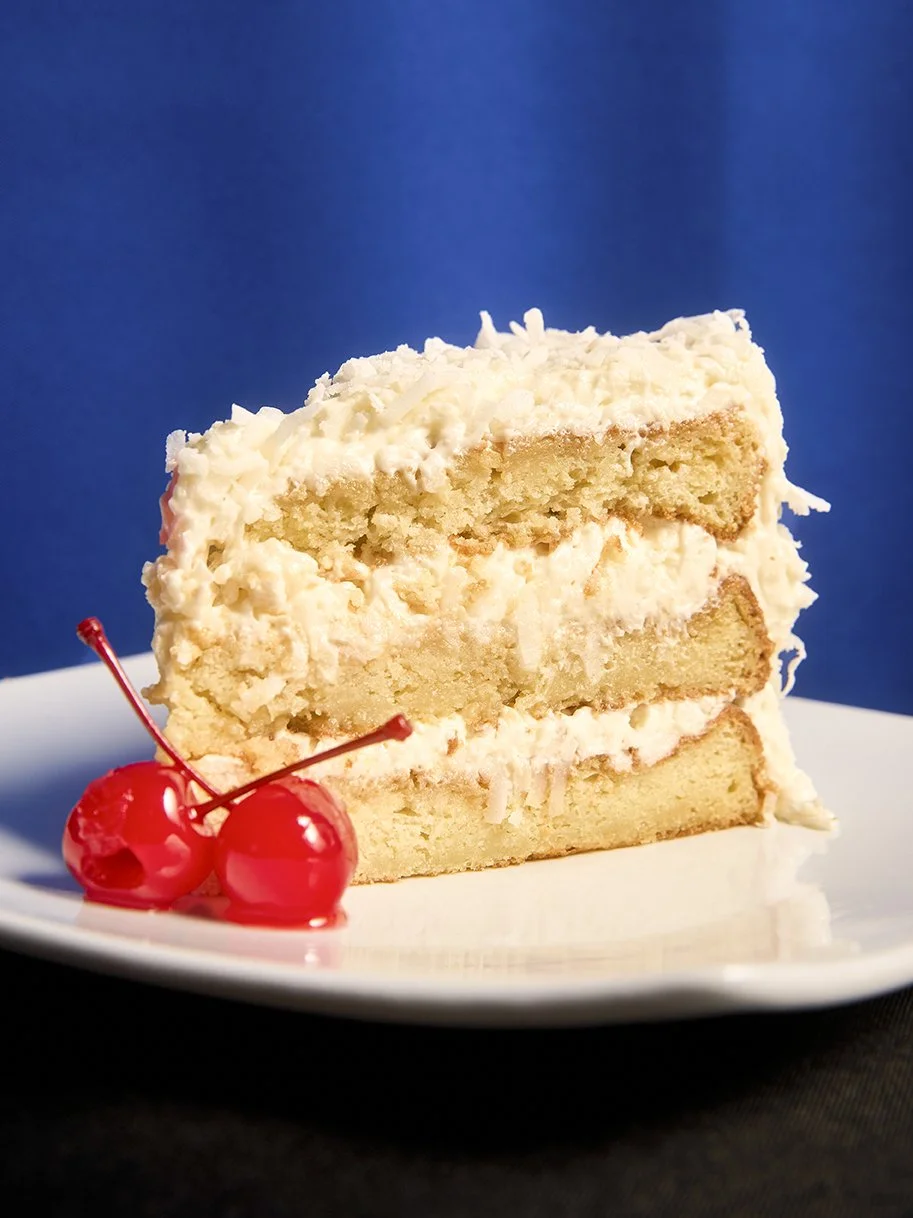 A slice of vanilla cake with white frosting on a white plate, garnished with two cherries, against a blue background.