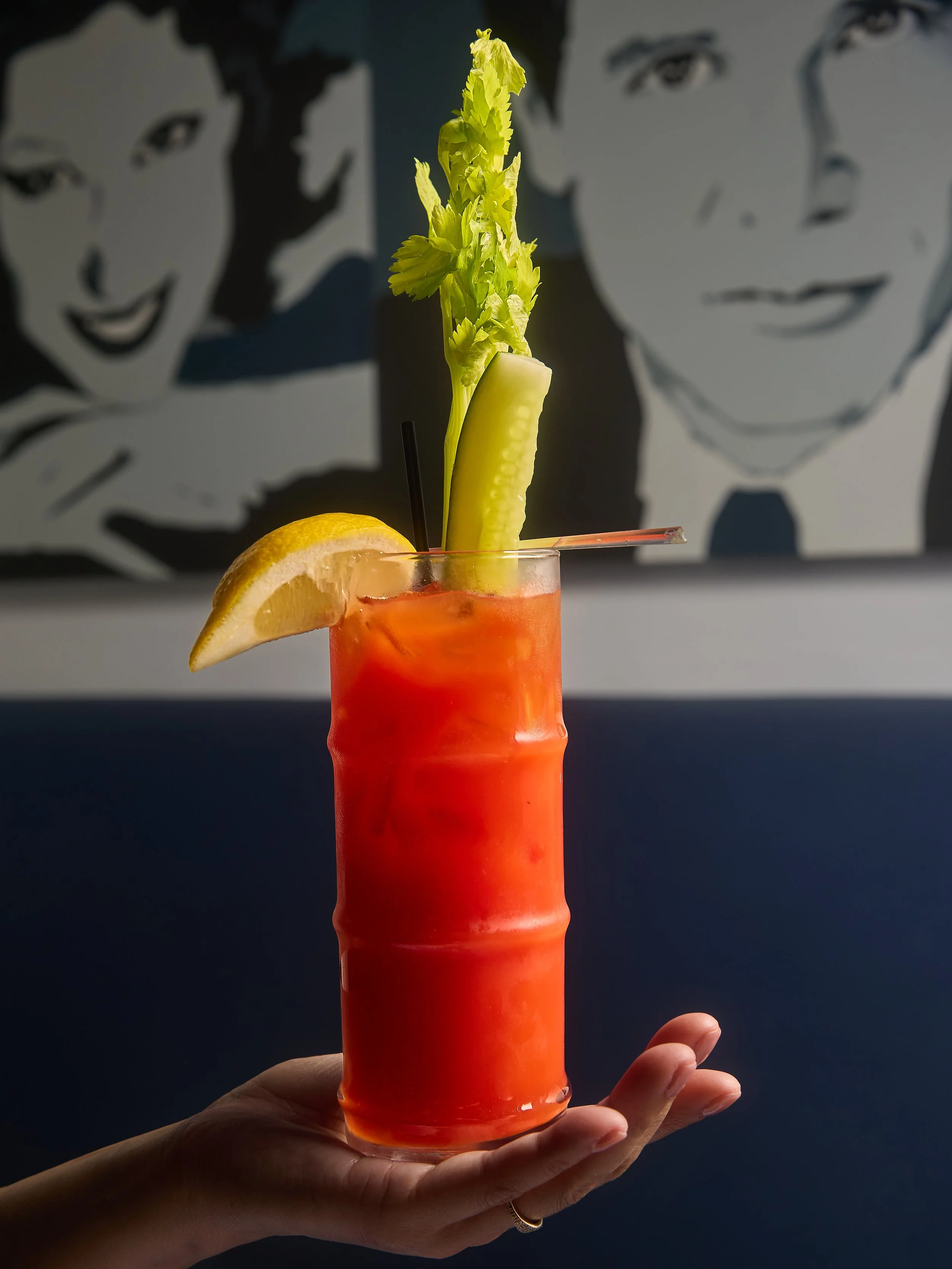 A hand holding a tall glass of pink cocktail with a lemon wedge, celery stalk, and straw, with a blurred black and white cartoon portrait in the background.