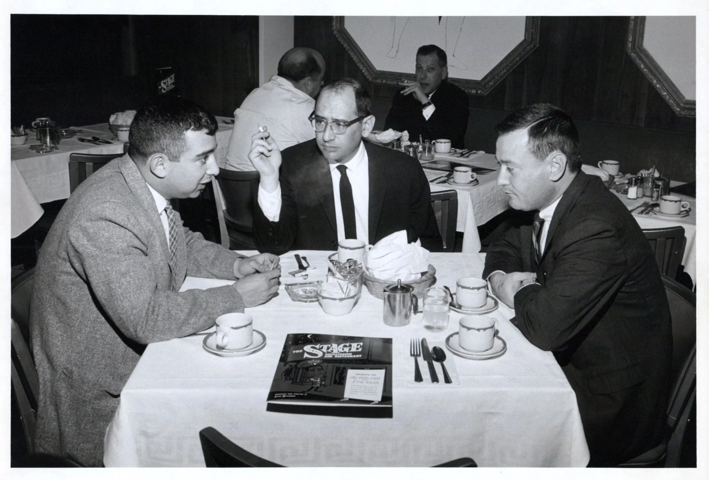 A black and white photograph of five men seated around a dining table in a restaurant, engaged in conversation. The table has cups, plates, and silverware, with a restaurant menu titled 'STAGE' in the foreground.