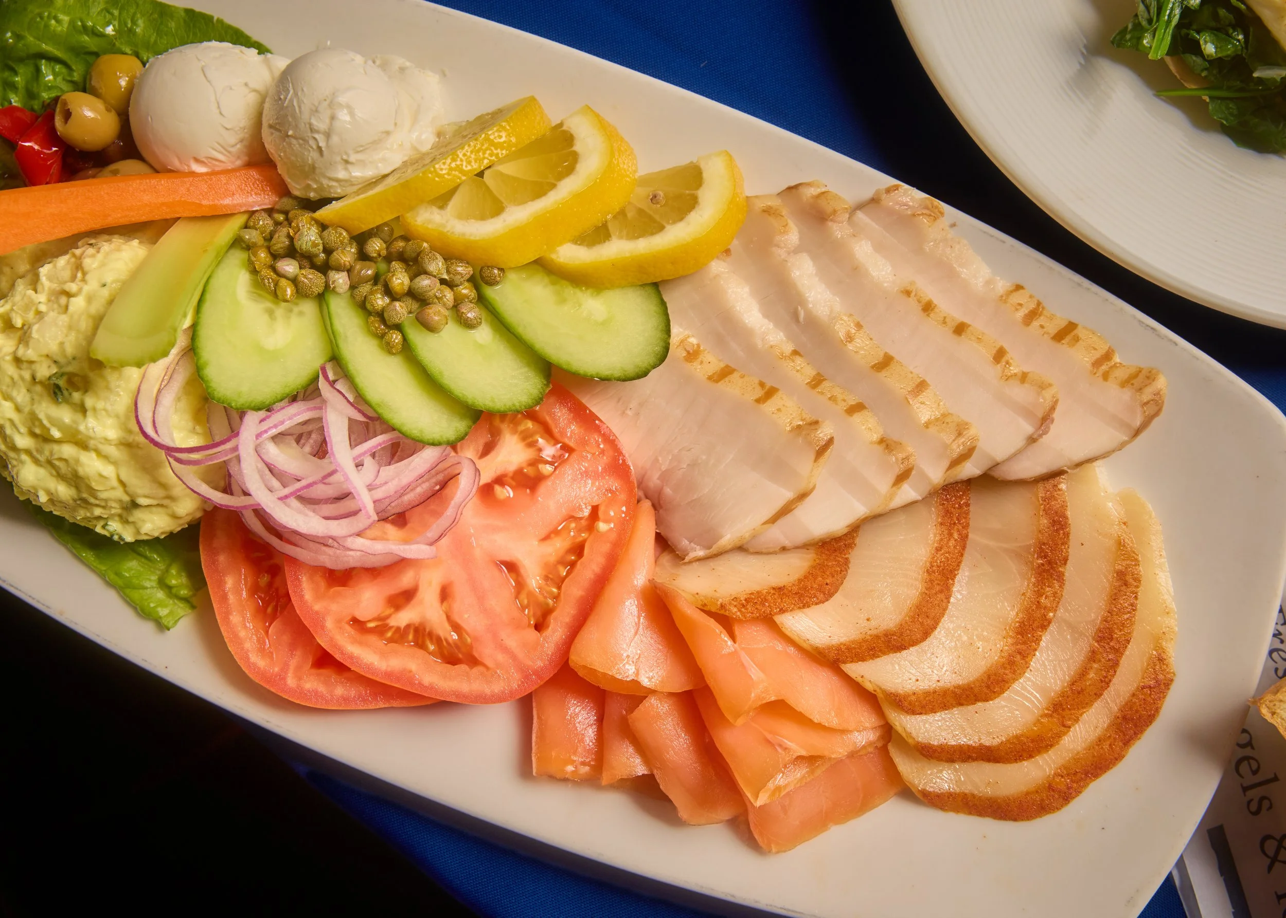 A white platter of assorted deli meats and fresh vegetables, including sliced turkey, smoked salmon, tomato slices, cucumber slices, lemon wedges, pickled onions, chickpeas, shredded lettuce, and sliced hard-boiled eggs.
