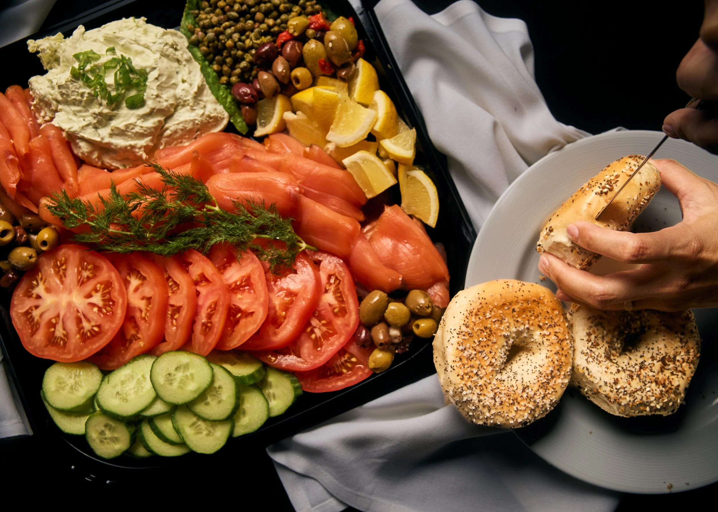 A platter of fresh foods including sliced tomatoes, sliced cucumbers, a dollop of potato salad garnished with green onions, a variety of green and black olives, and lemon wedges. There are also two bagels topped with seeds, one with a person slicing it with a knife.