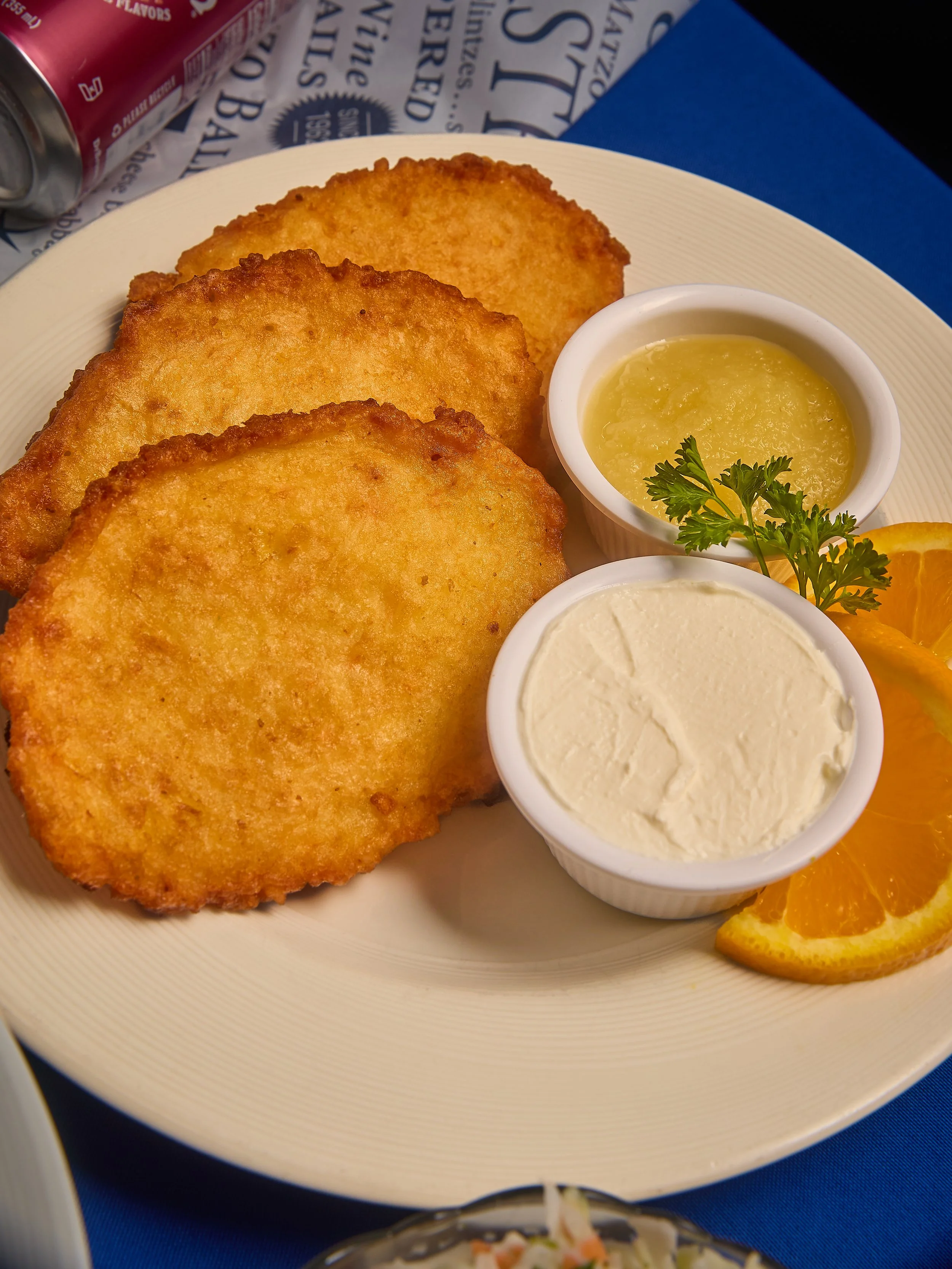 Three golden-brown fried fish fillets on a white plate with two small cups of tartar sauce and lemon wedges garnished with parsley, with a blue tablecloth underneath.