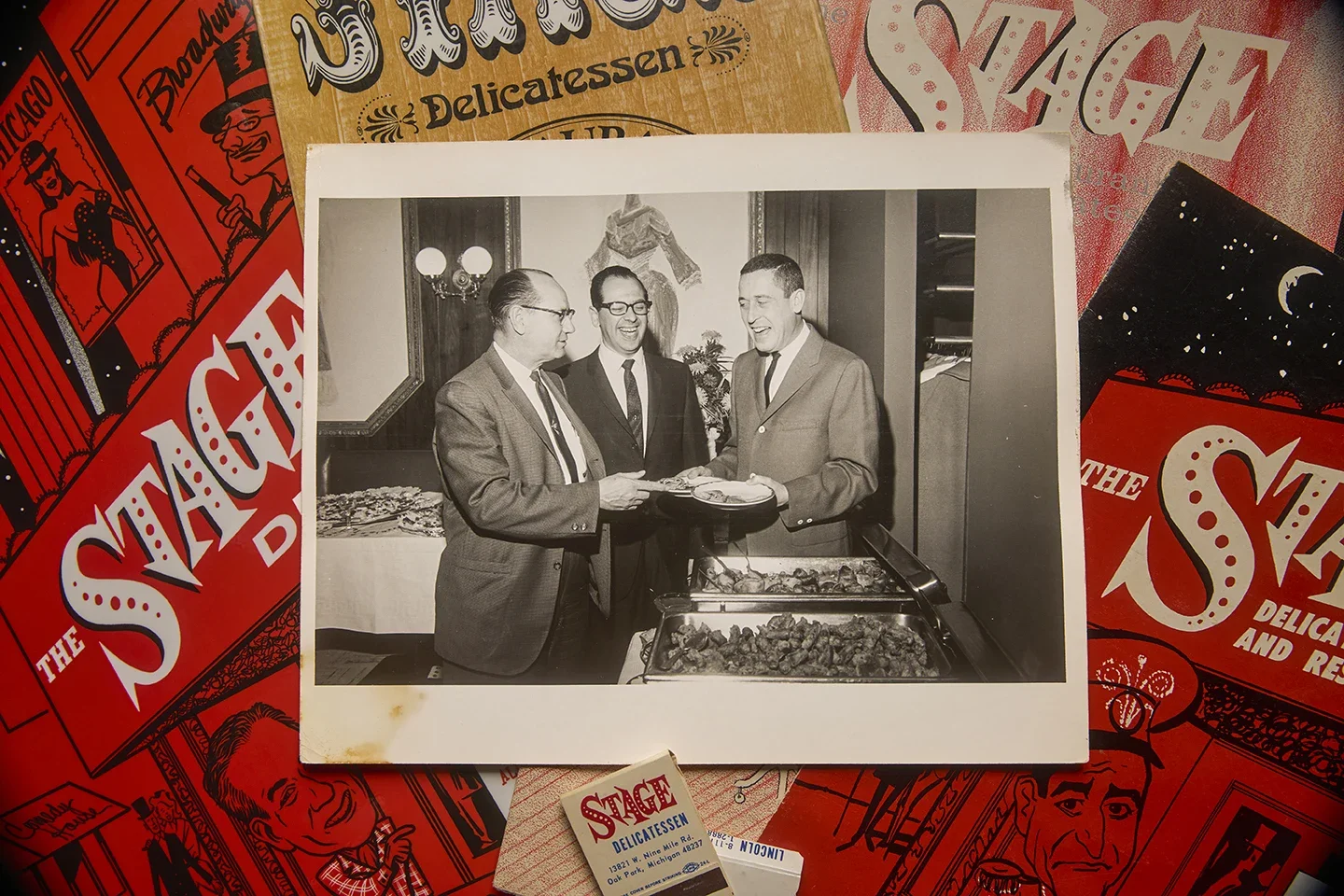 Black and white photograph of three men in suits smiling and talking at a buffet table, with food trays in front of them.
