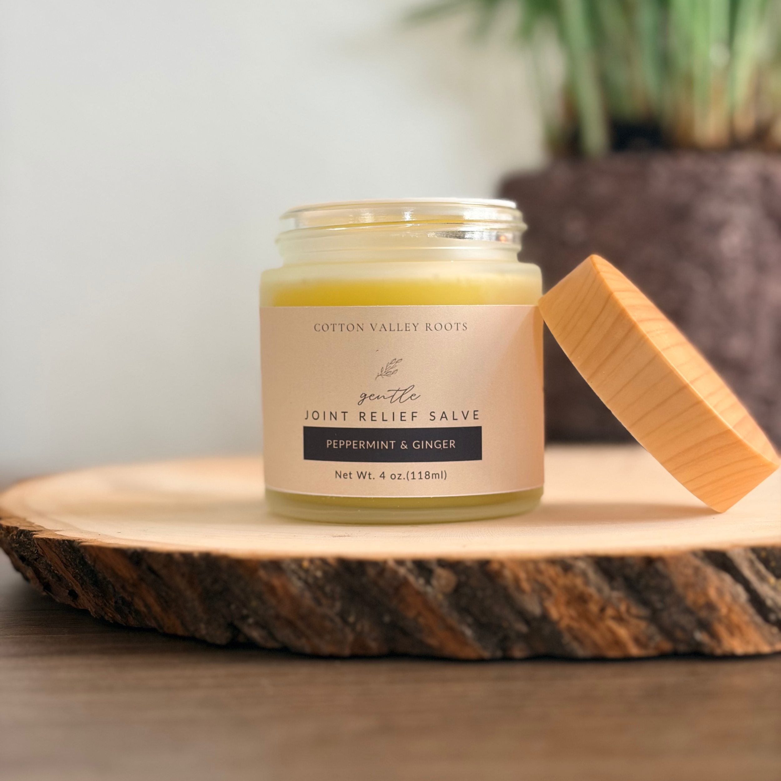 Close-up of a jar of Cotton Valley Roots joint relief salve with peppermint and ginger, placed on a wooden surface with a blurred plant in the background.