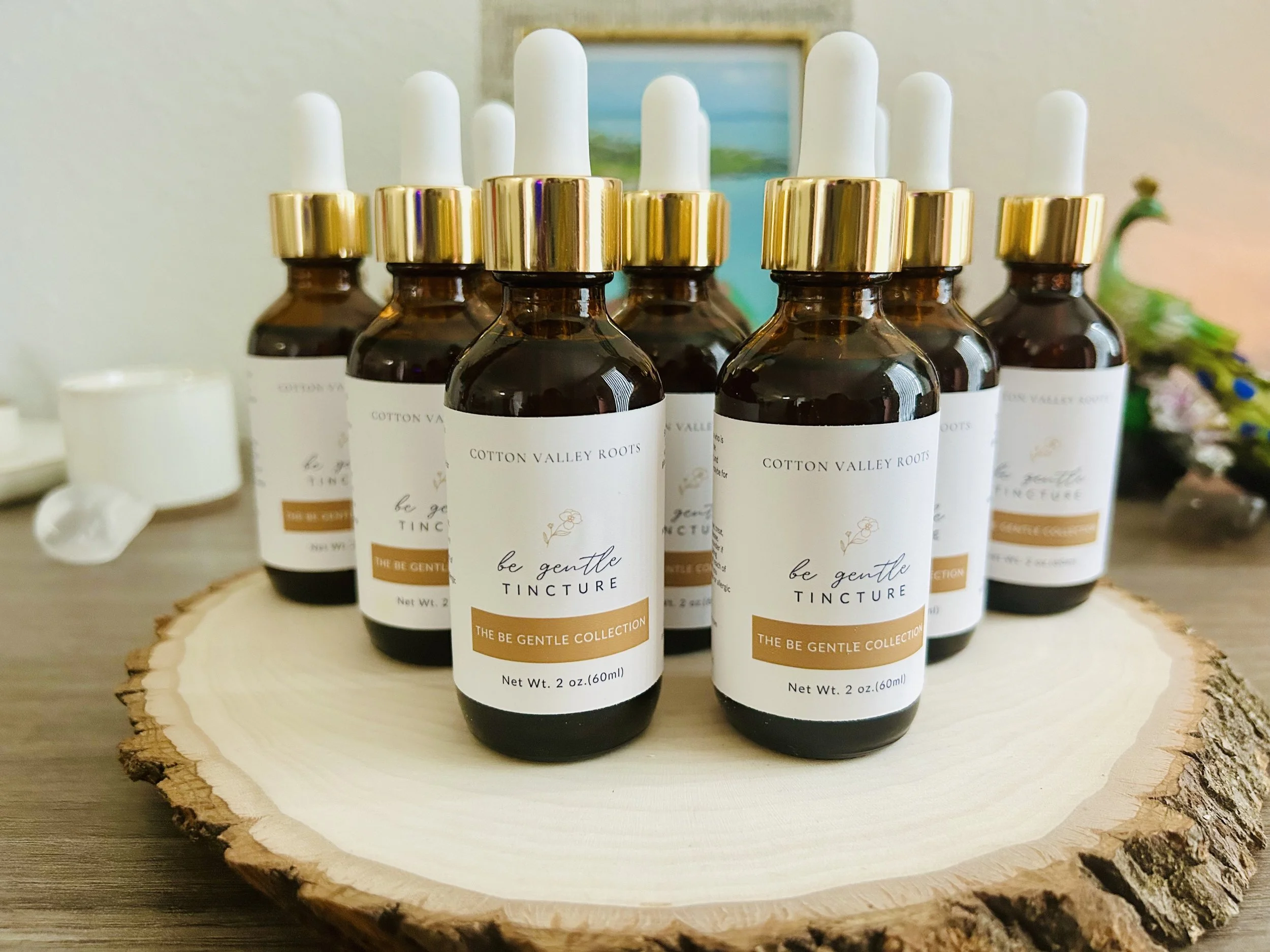Several amber glass dropper bottles with white caps and labels reading "be gentle TINCTURE" arranged on a wooden slice with a blurred background.