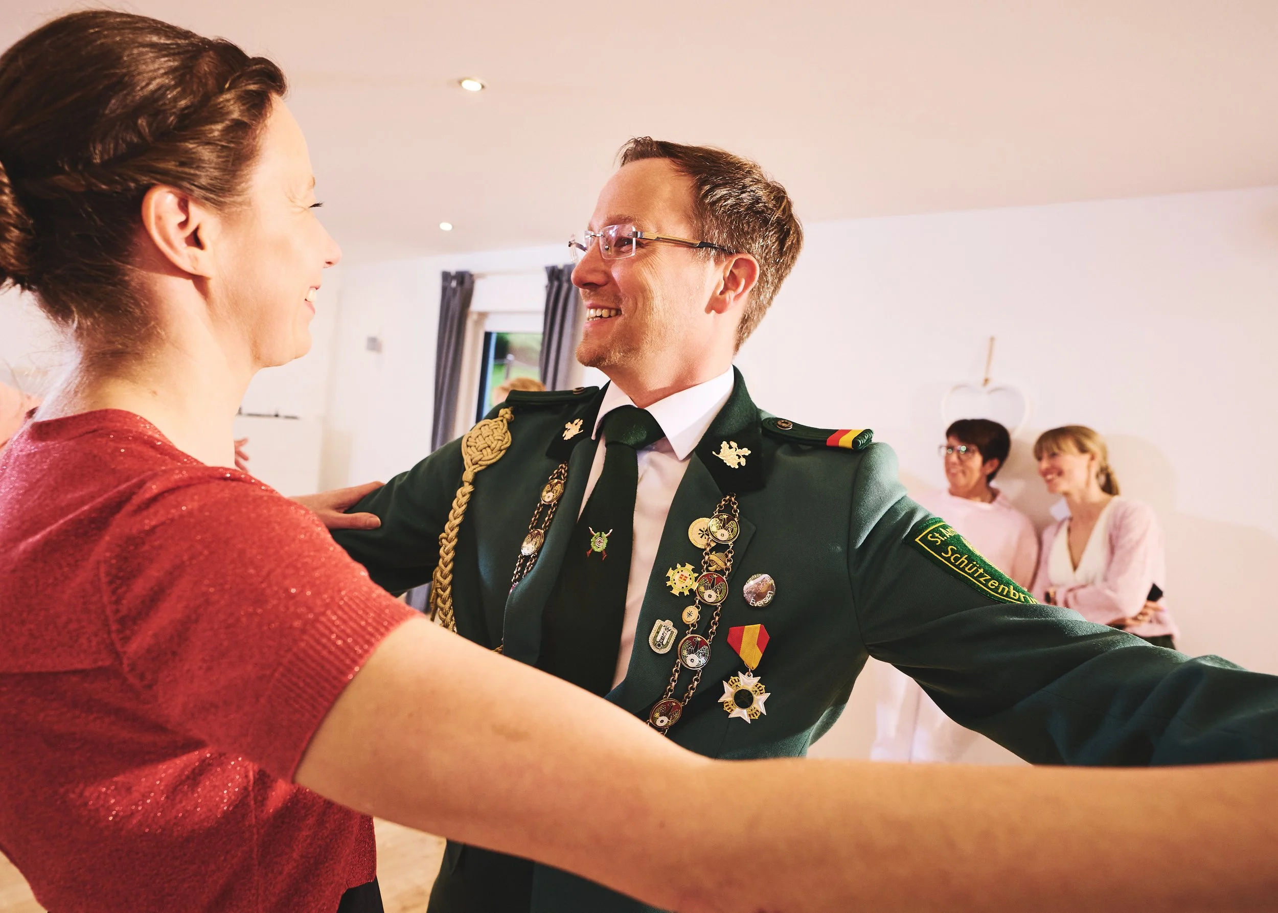 In der Tanzschule Winkler.  Eine Frau in einem roten Kleid und ein Mann in einer grünen Uniform tanzen miteinander. Im Hintergrund sind weitere Frauen, die ebenfalls tanzen, und die Szenerie ist in einem hellen Raum mit Vorhängen und Deckenleuchten.