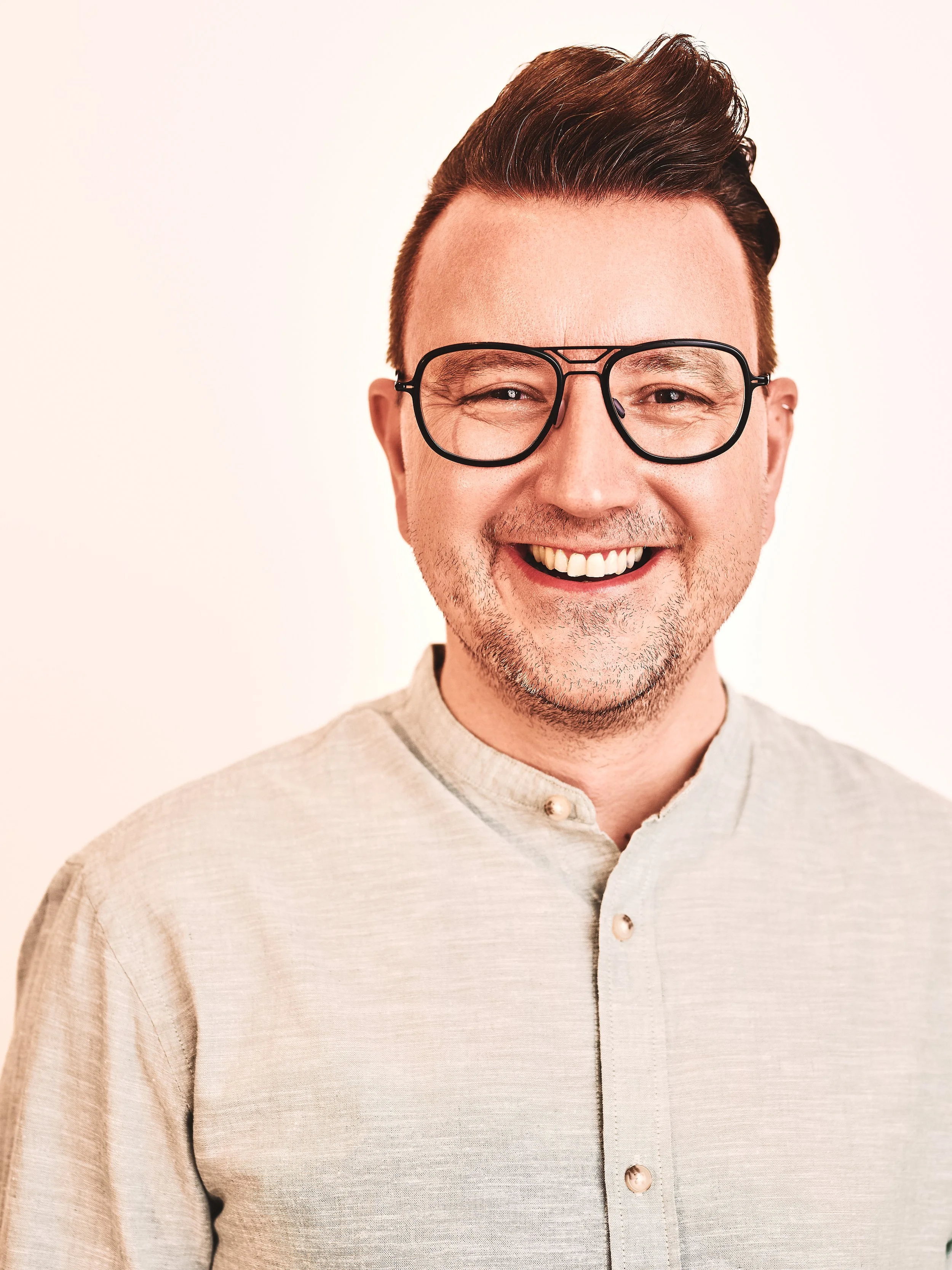 Ein Mann mit braunen Haaren, Brille und einem cremefarbenen Hemd lächelt. Hintergrund ist weiß.