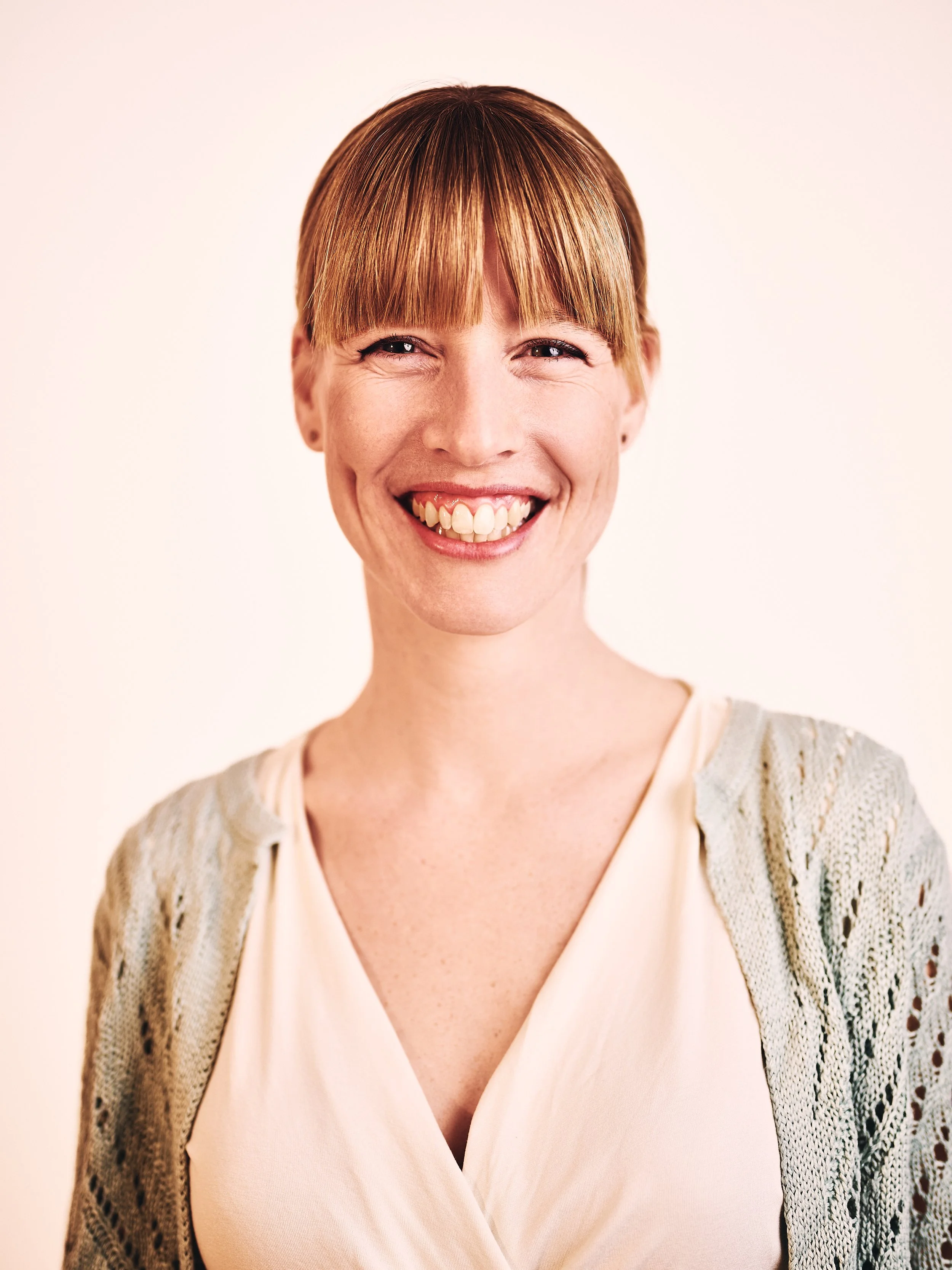 Fröhliche Frau mit rotem Haar und Zopf, trägt Cremefarbene Bluse und einen Strickcardigan, lachend vor weißem Hintergrund.