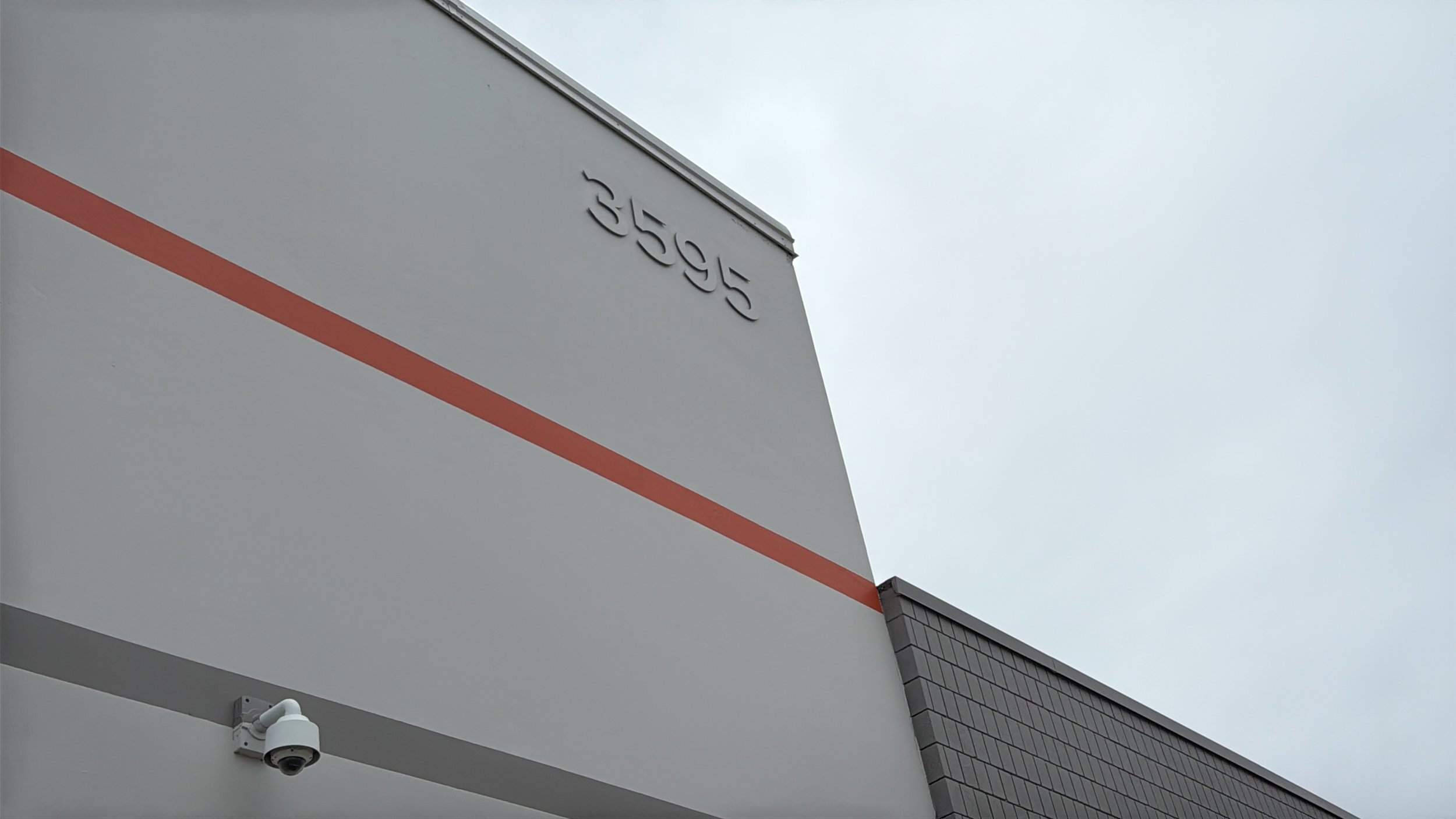 Close-up of a modern building exterior with a surveillance camera and the number 355695 displayed on the corner, against an overcast sky.