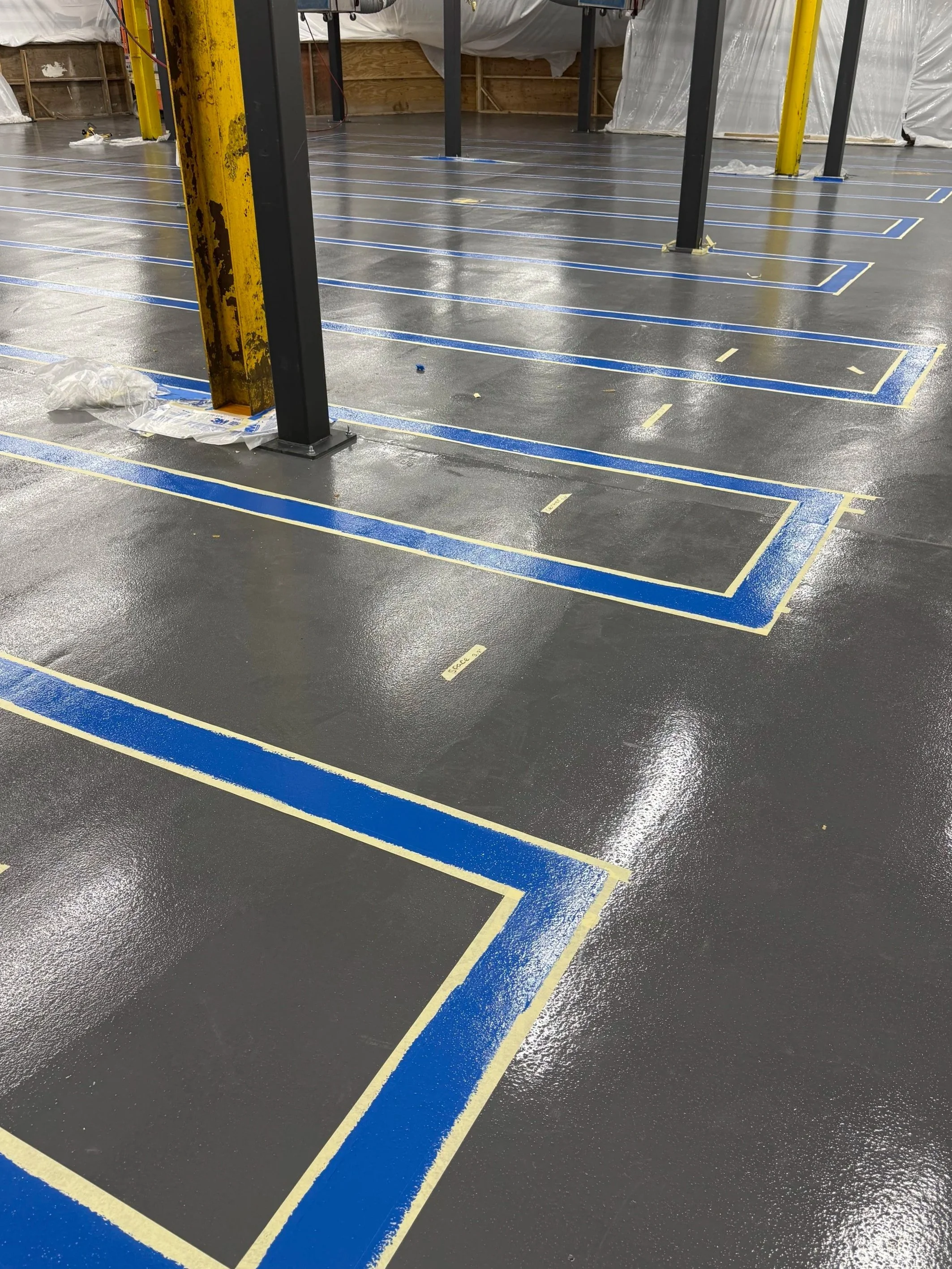 An indoor construction site with a polished concrete floor, marked with blue and yellow painter’s tape forming various rectangular and linear patterns. There are black metal support beams, one rusty yellow beam, and plastic sheeting covering parts of the wall.
