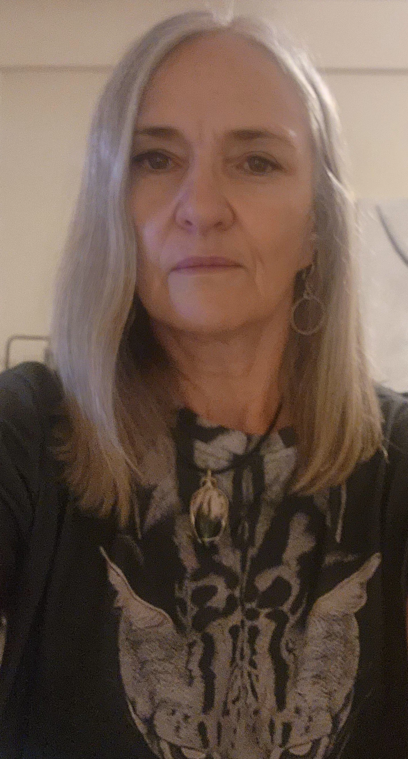 A woman with shoulder-length blonde hair wearing a black and beige animal print top and earrings, against a beige wall.