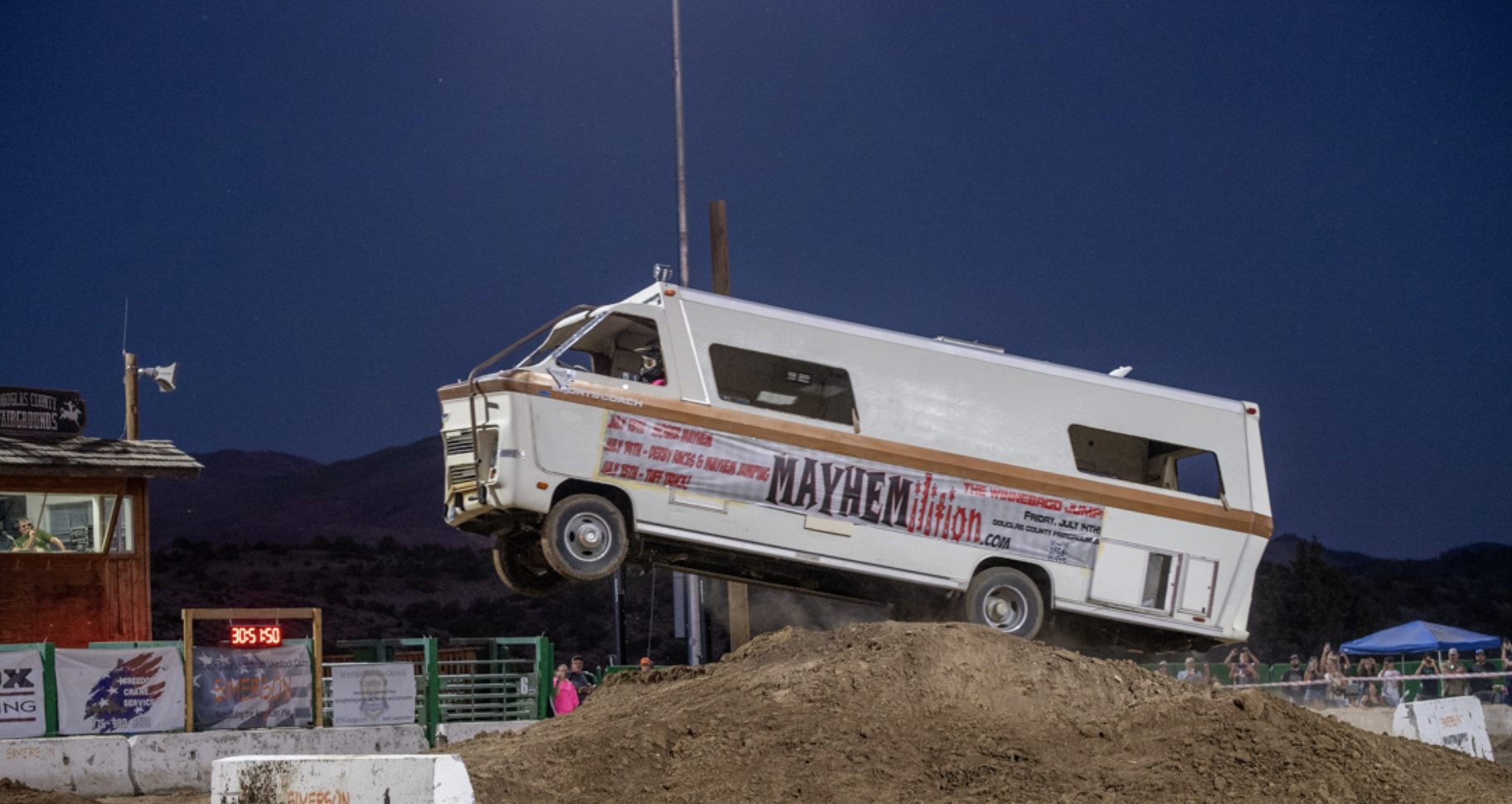 A vintage camper van in mid-air during a stunt at a dirt track event at night, with spectators in the background and banners nearby.