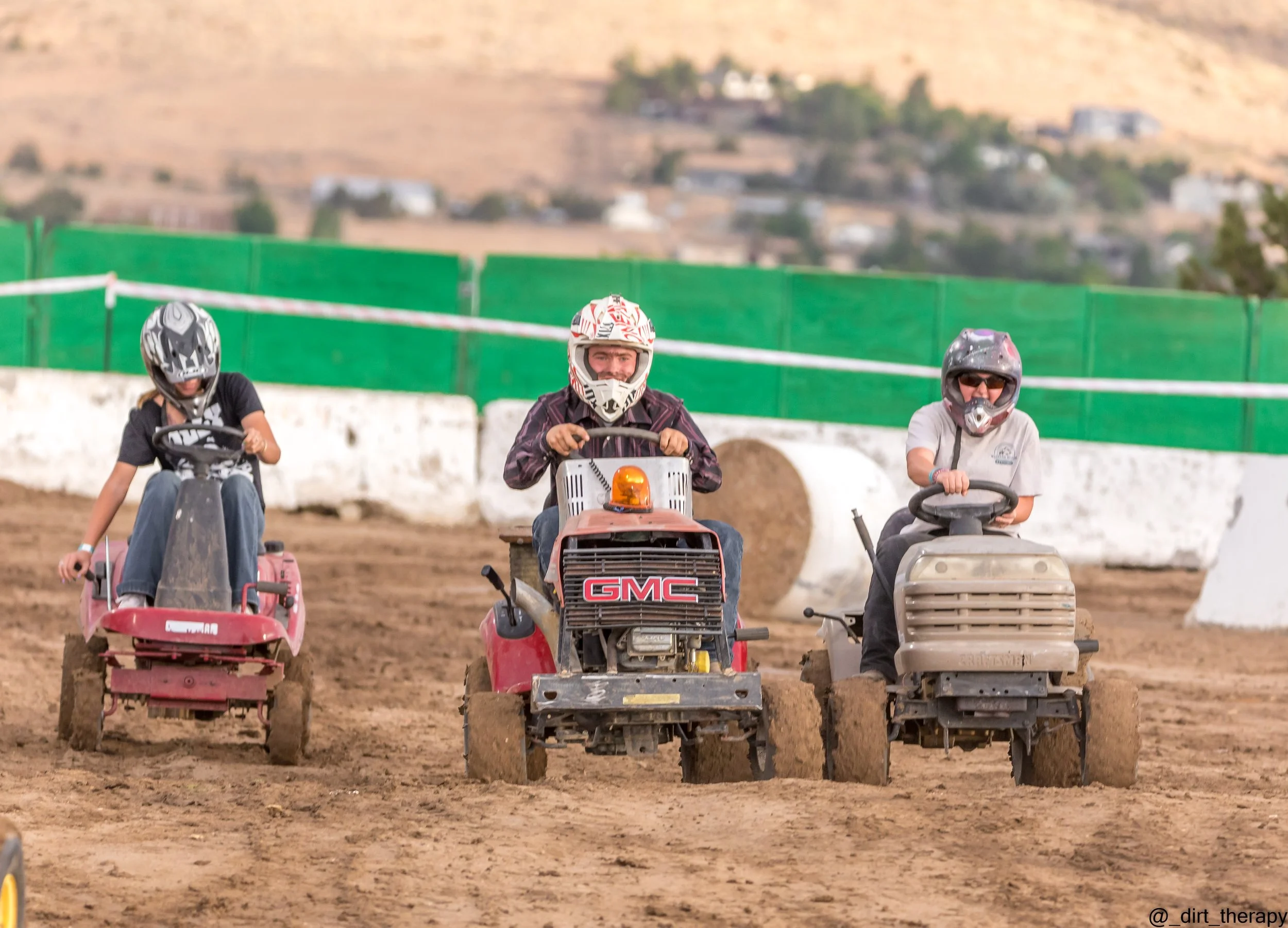 Three people wearing helmets riding small riding lawn mowers on a dirt track, with a green fence and houses on a hillside in the background.