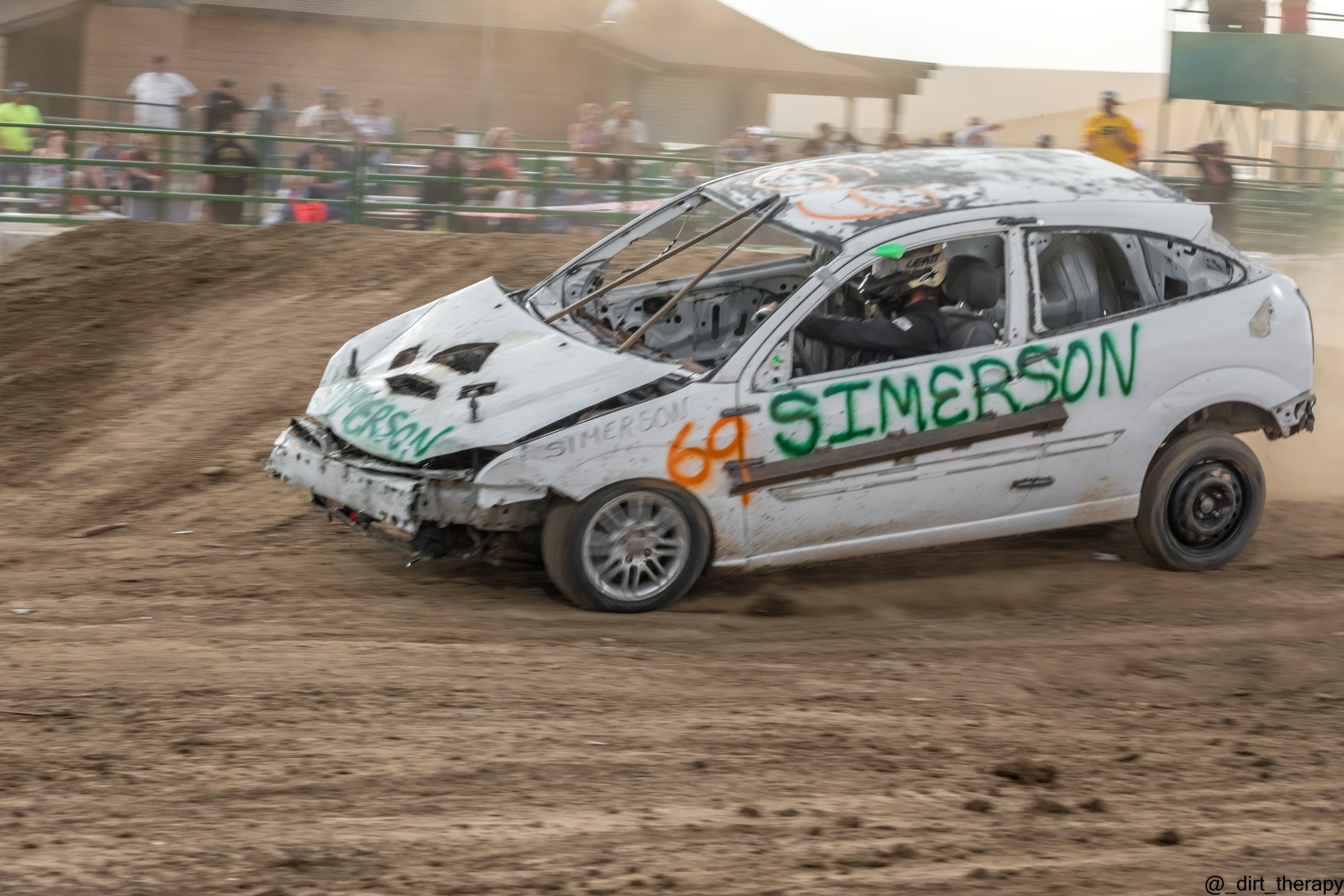 A damaged race car on a dirt track with spectators watching in the background.
