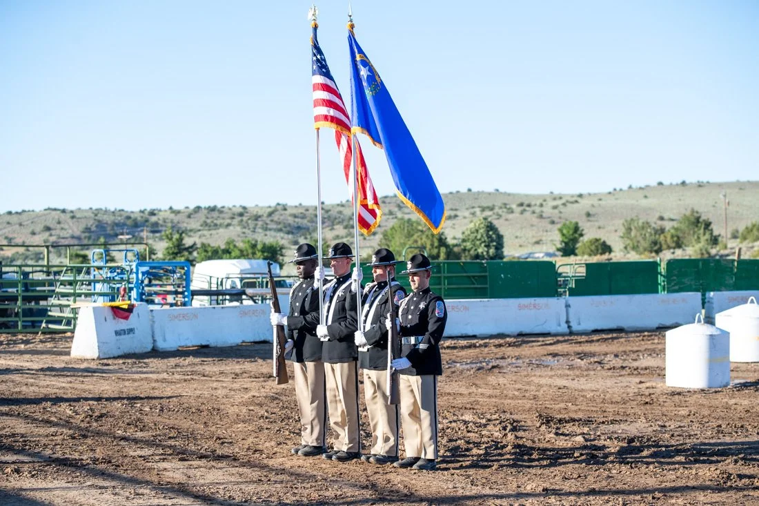 Four uniformed men standing in formation holding flags, including the American flag, on a dirt field with hills in the background.