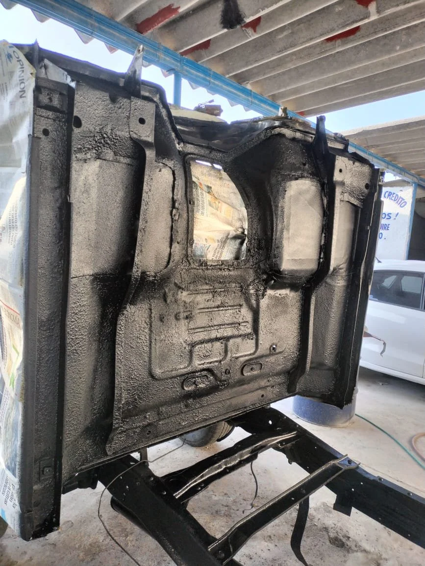 Chasis de vehículo pintado de negro en reparación, en un taller con estructura de techo de madera y coche blanco visible en el fondo.